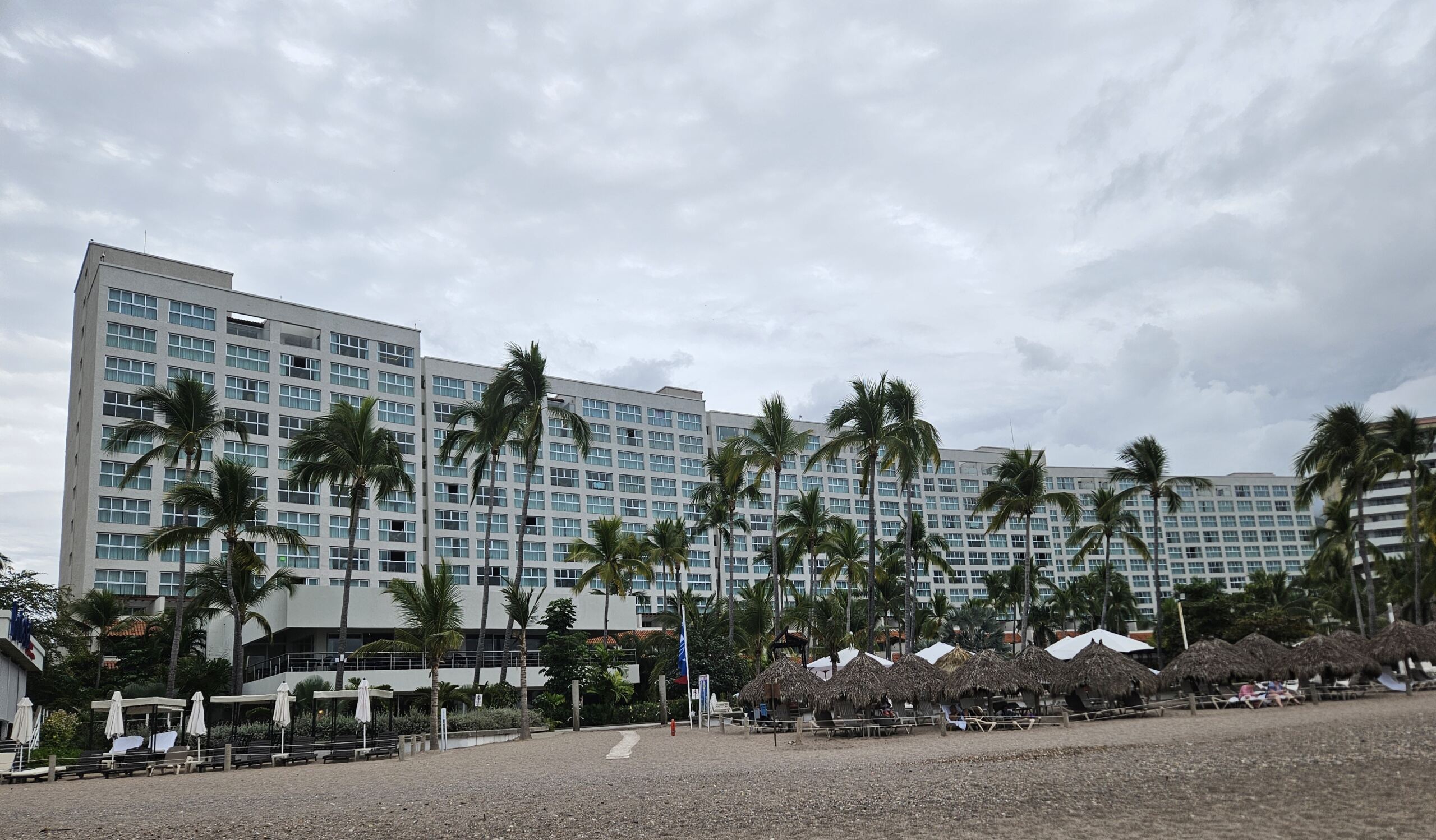 Ein Strandhotel mit einem hohen weißen Gebäude und zahlreichen Fenstern steht stolz da und erinnert an einen Veranstaltungsort, an dem prestigeträchtige Veranstaltungen wie die Preisverleihung des Best Paper Award stattfinden. Palmen säumen den Sandstrand, unter einem bewölkten Himmel stehen Sonnenliegen und Sonnenschirme. at Hochschule Coburg