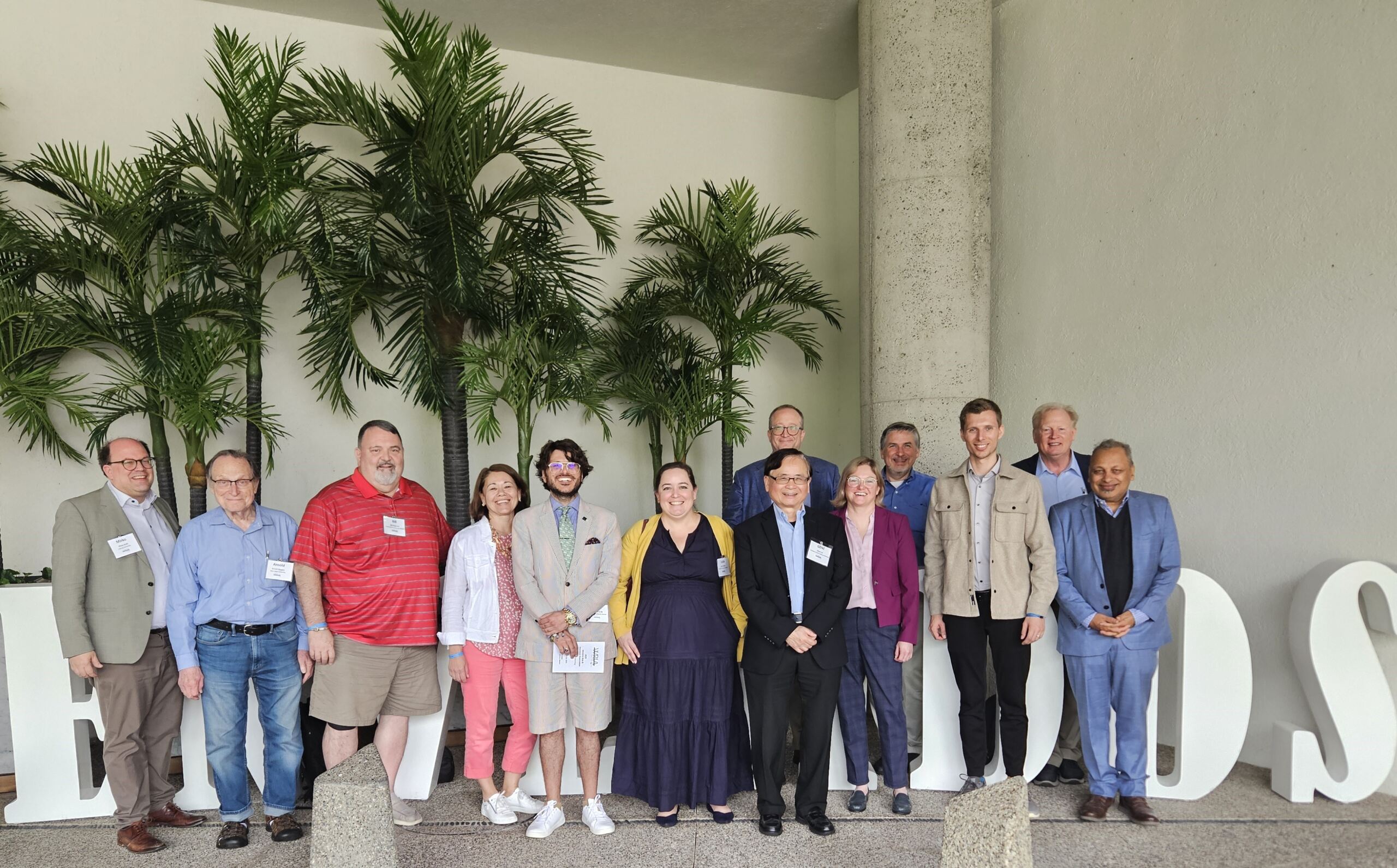 Eine bunt gemischte Gruppe von Menschen steht in einer Reihe und lächelt in die Kamera auf der US-Konferenz. Sie stehen vor großen Buchstaben und sind von üppigen grünen Pflanzen umgeben. Die Personen, darunter ein Gewinner des Best Paper Award, tragen eine Mischung aus formeller und legerer Kleidung. at Hochschule Coburg