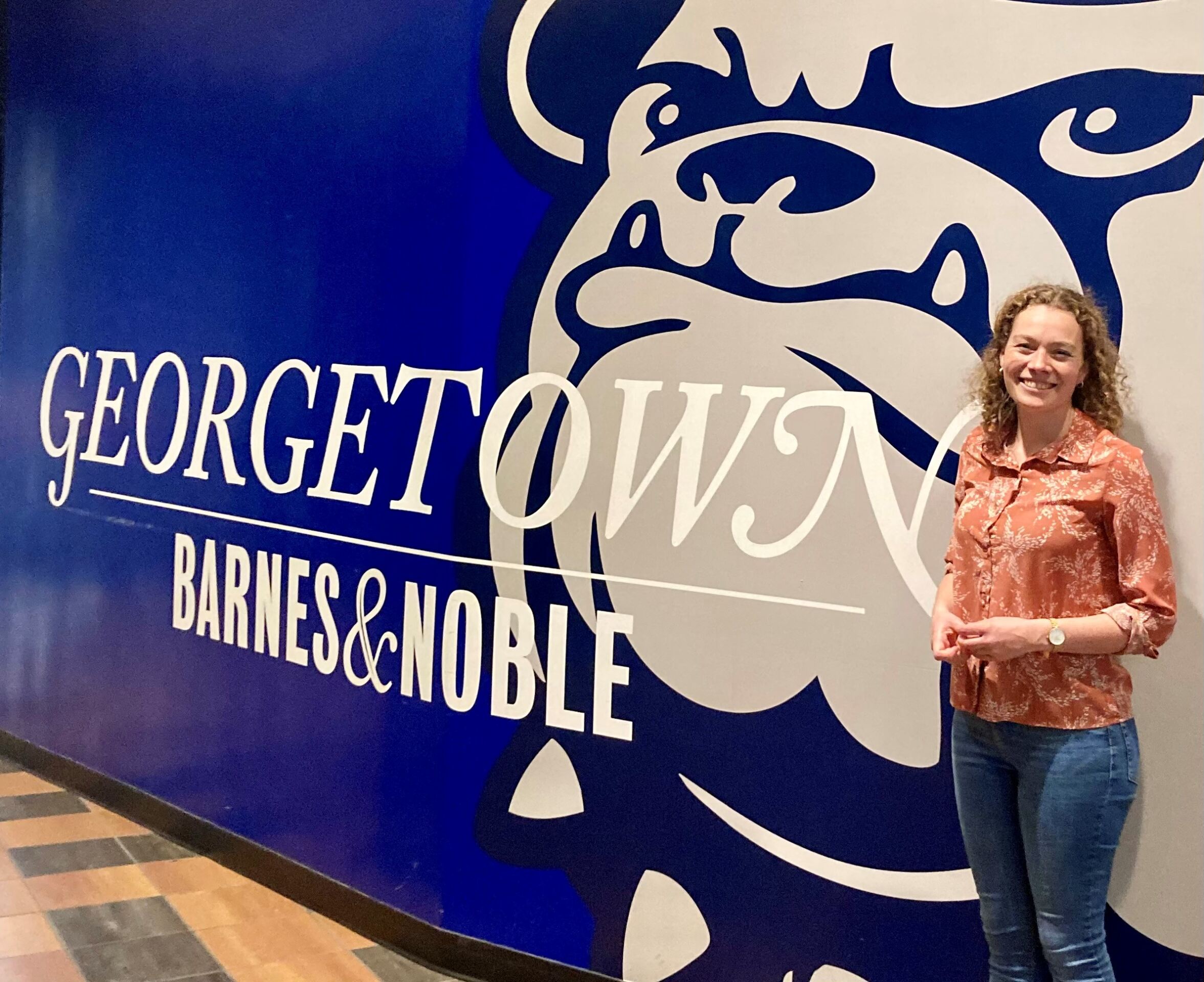 Eine Person steht lächelnd neben einer Wand mit einer großen Bulldoggengrafik und dem Text „Georgetown Barnes & Noble“. Der Hintergrund ist blau und weiß und die Person trägt eine gemusterte orangefarbene Bluse und Jeans. at Hochschule Coburg