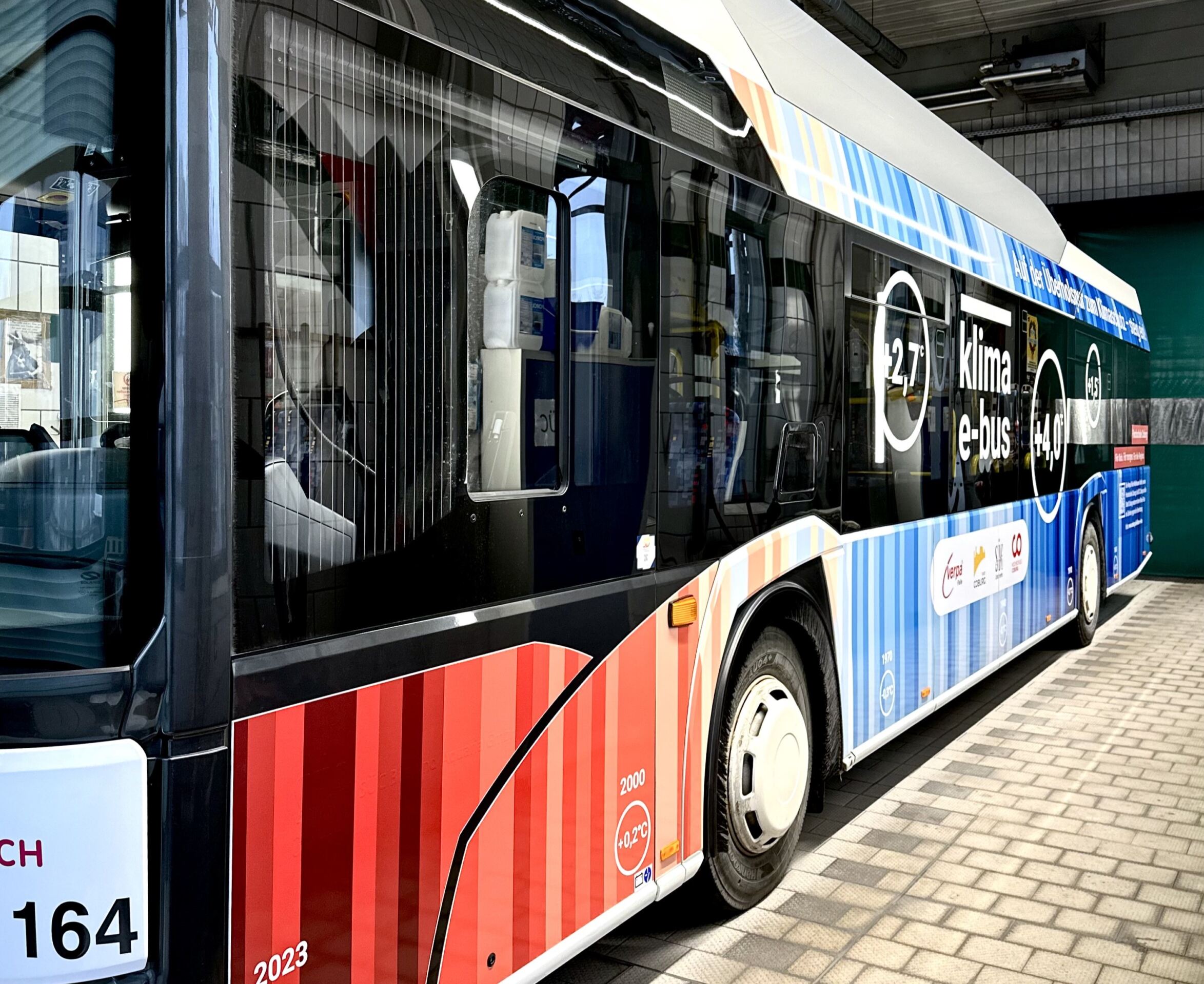 Ein farbenfroher Elektrobus steht in einer Garage. Der Bus hat ein Streifendesign in Rot- und Blautönen und zeigt Texte und Logos zu den Themen Klima und Energieeffizienz. Die Zahl 27 ist deutlich zu sehen. at Hochschule Coburg