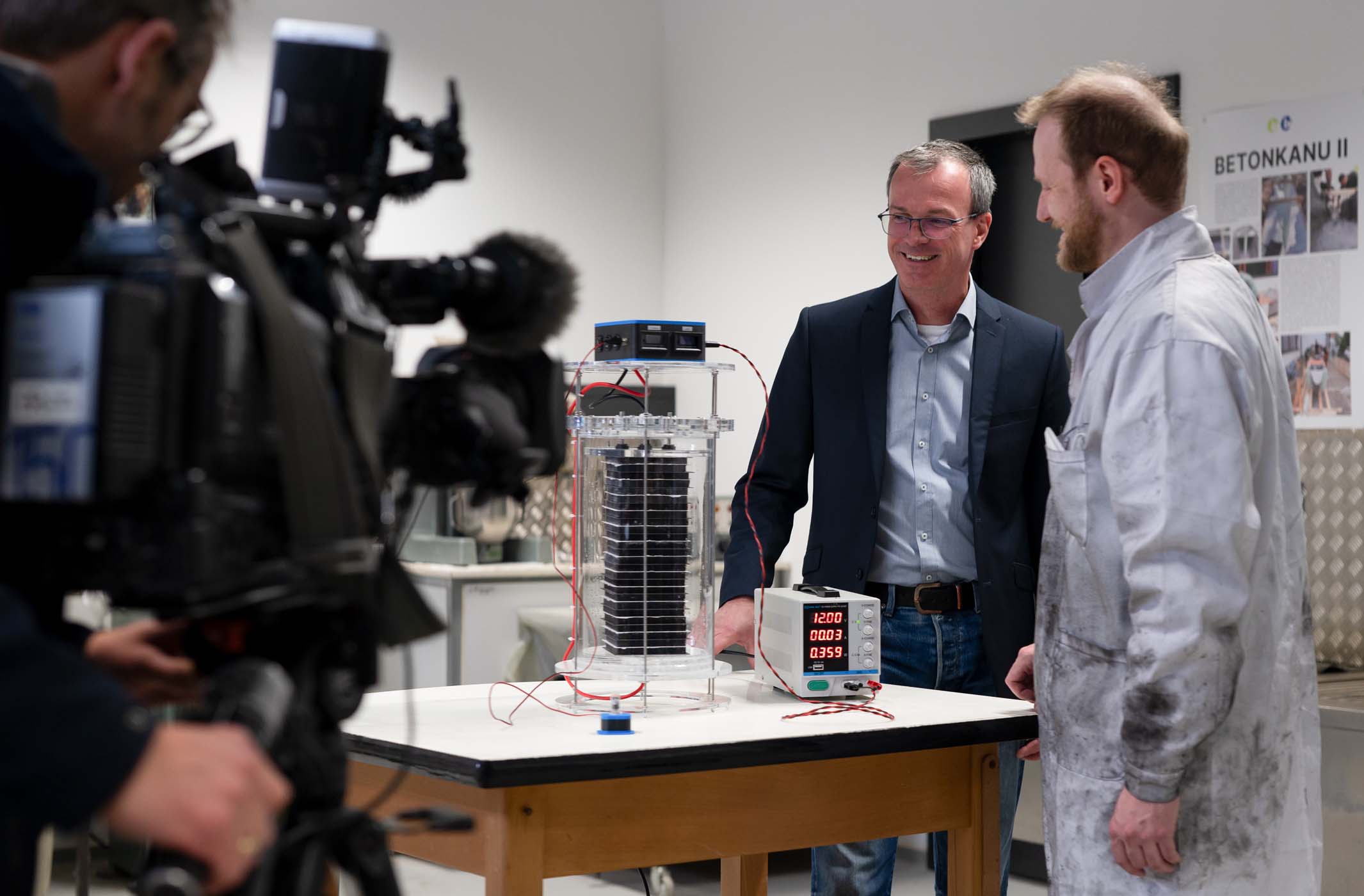 Zwei Männer stehen neben einem wissenschaftlichen Gerät auf einem Tisch und unterhalten sich. Der eine trägt einen Anzug, der andere einen Laborkittel. Ein Kamerateam filmt sie, während sie über den Stromspeicher diskutieren, umgeben von Kabeln und einem digitalen Bildschirm mit Zahlen. at Hochschule Coburg