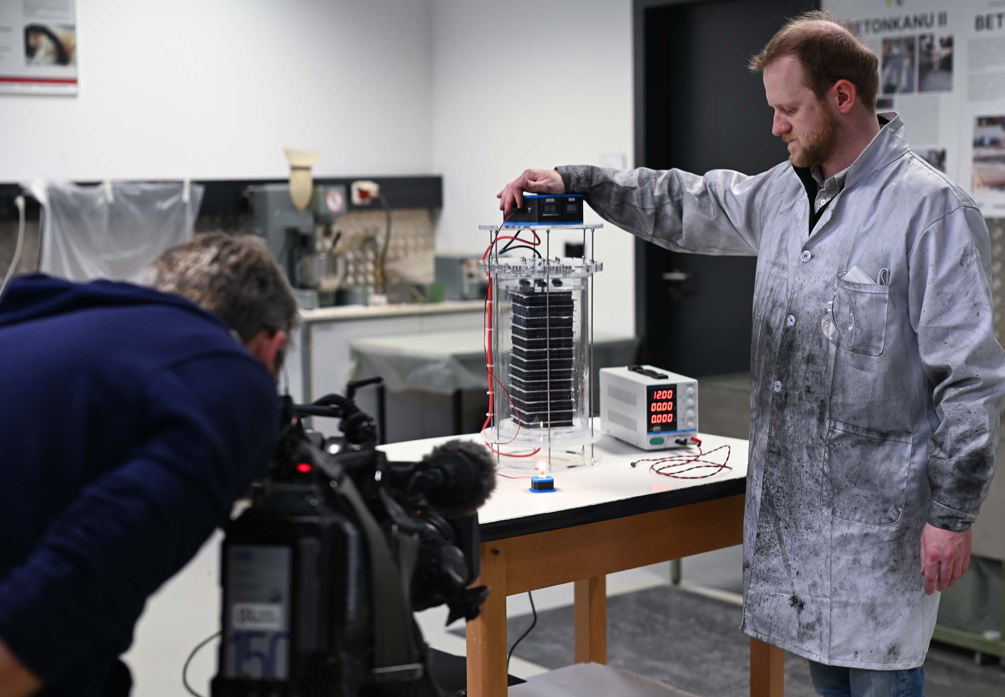 Ein Mann im Laborkittel führt ein Experiment mit elektrischen Geräten durch. Dabei konzentriert er sich auf einen Stromspeicher, der mit Drähten an einen Zylinder angeschlossen ist. In der Nähe bedient eine Person eine Videokamera, um den Vorgang im Labor zu dokumentieren, das mit verschiedenen Werkzeugen und Postern gefüllt ist. at Hochschule Coburg