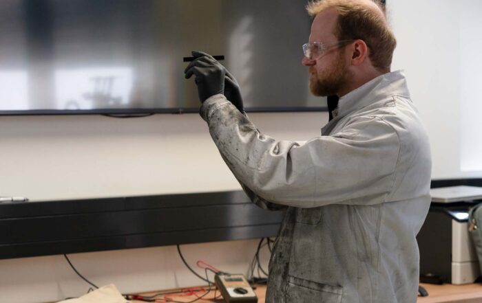 Eine Person in schmutzigem Laborkittel und Schutzbrille hält einen Stromspeicher hoch und untersucht ihn sorgfältig. Sie trägt schwarze Handschuhe. Im Hintergrund sind Laborgeräte und ein Flachbildschirm zu sehen. at Hochschule Coburg