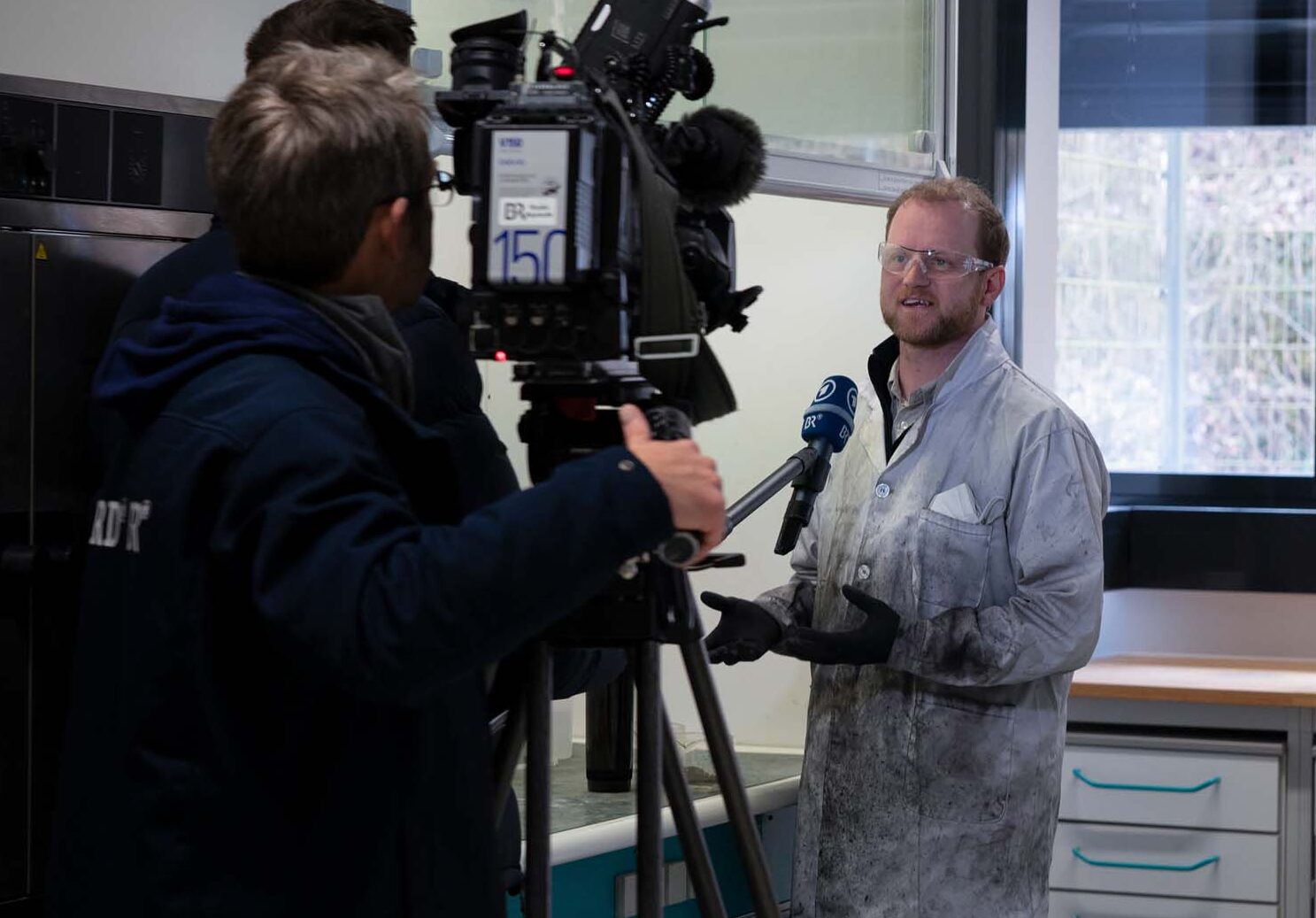 Eine Person im Laborkittel steht in einem Labor und spricht über Stromspeichertechnologie, während ein Kamerateam ihre Präsentation aufzeichnet. Vor ihr sind Mikrofone positioniert. Der Raum ist voller Geräte, und ein Fenster mit teilweise geöffneten Jalousien wirft Lichtstreifen hinein. at Hochschule Coburg