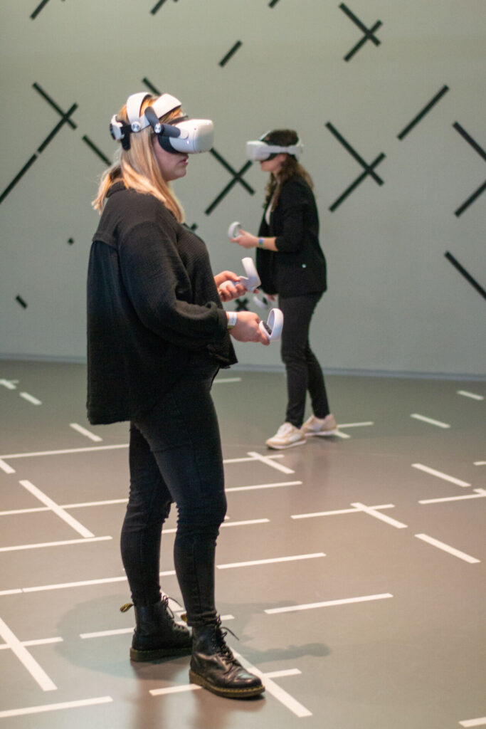 Zwei Personen in einem Raum tragen Virtual Reality-Headsets und halten Controller in der Hand. Sie stehen auf einem mit schwarzen und weißen Linien markierten Boden. Die Wände sind mit schwarzen diagonalen Linien verziert. Beide Personen sind schwarz gekleidet. at Hochschule Coburg