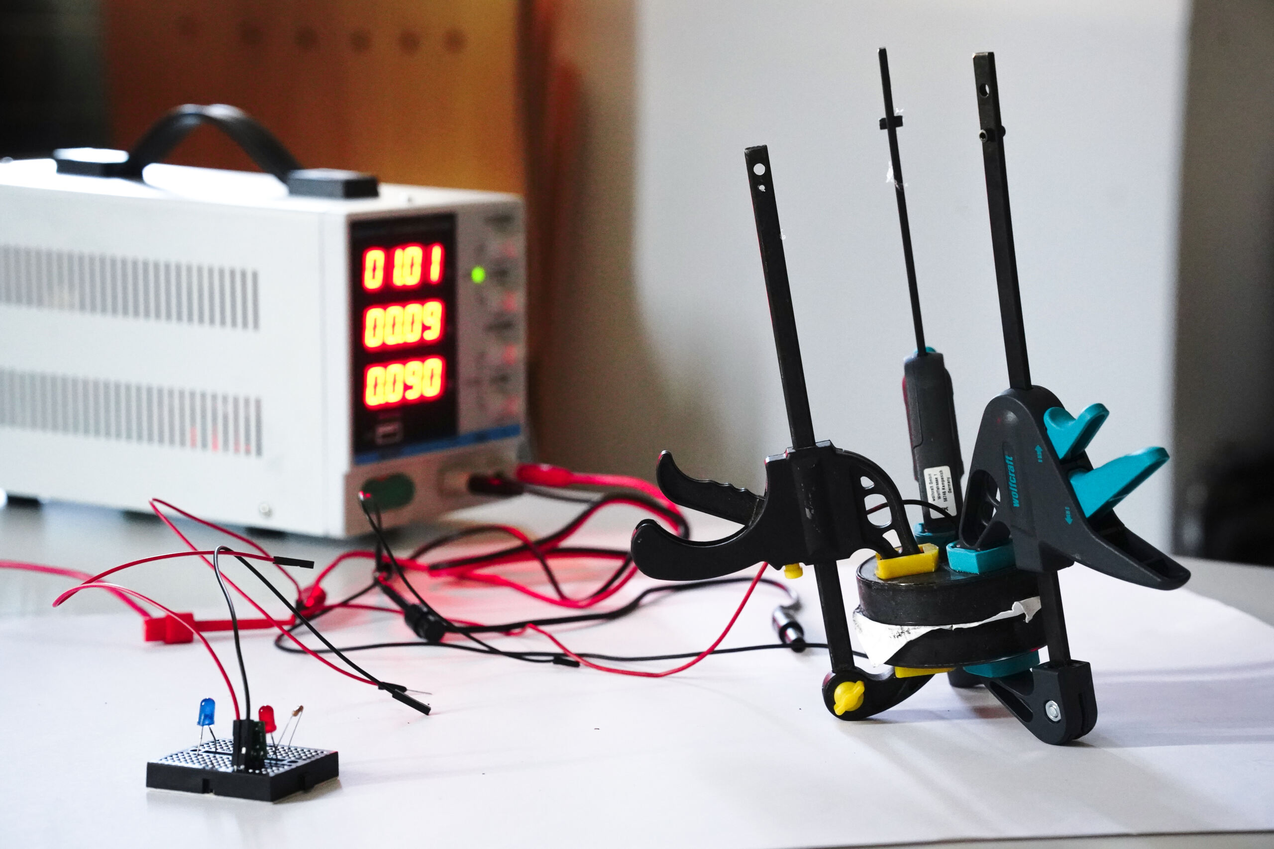 Ein Netzteil mit einer roten Digitalanzeige ist an einen Schaltkreis angeschlossen, der einem Stromspeicher ähnelt. Der Aufbau umfasst Kabel und ein Steckbrett mit blauen und roten LEDs. In der Nähe liegen Klemmen und ein Schraubendreher auf der festen Oberfläche, die an Betonstabilität erinnert. at Hochschule Coburg