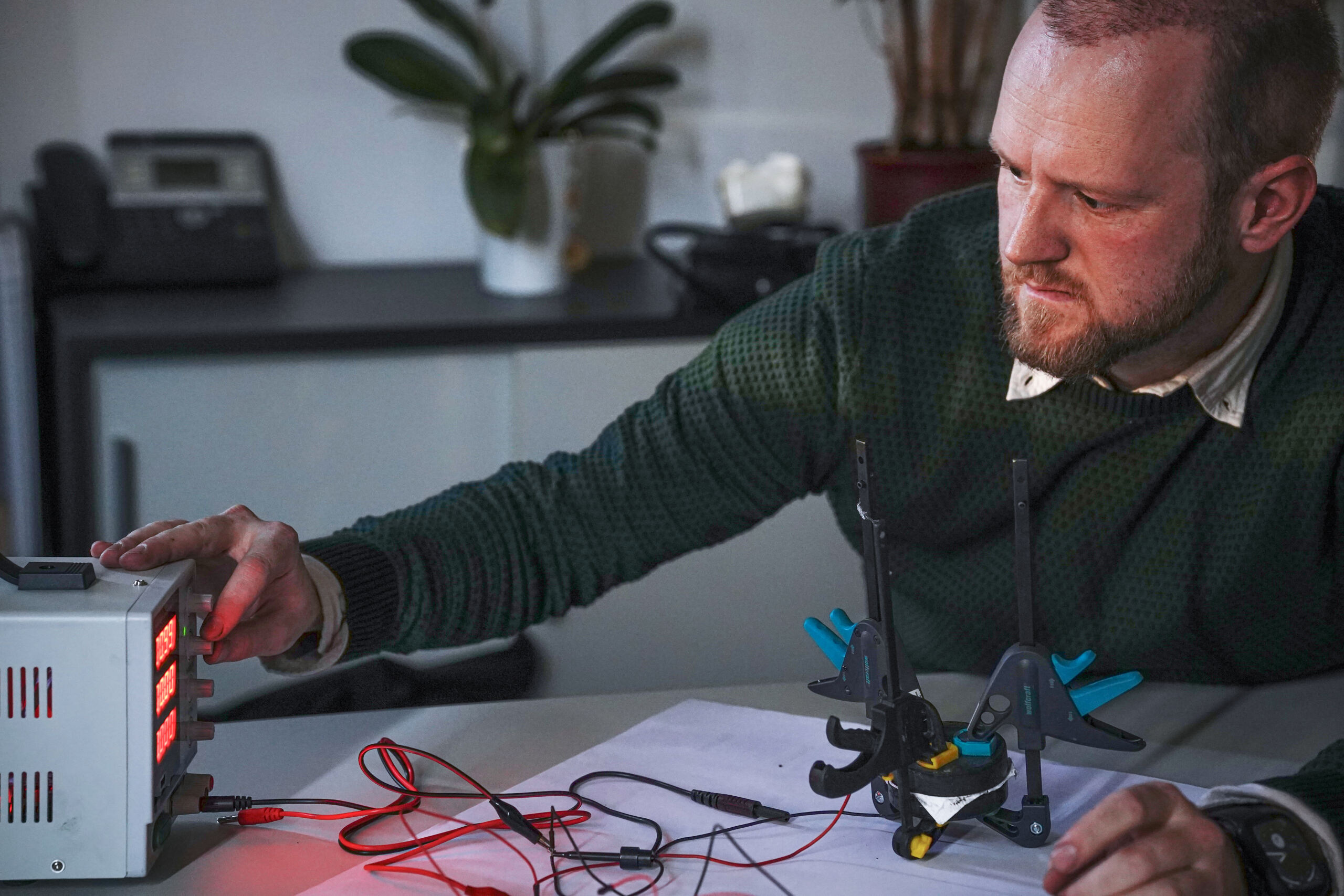 Ein Mann justiert eine Steuerbox, die über Kabel mit einem kleinen Gerät auf dem Tisch verbunden ist. Das Gerät, sicher mit Klammern gehalten, ruht auf einem Blatt Papier mit skizzierten Diagrammen, möglicherweise für ein Stromspeicherprojekt. Im Hintergrund sind Anlagen und Bürogeräte zu sehen. at Hochschule Coburg