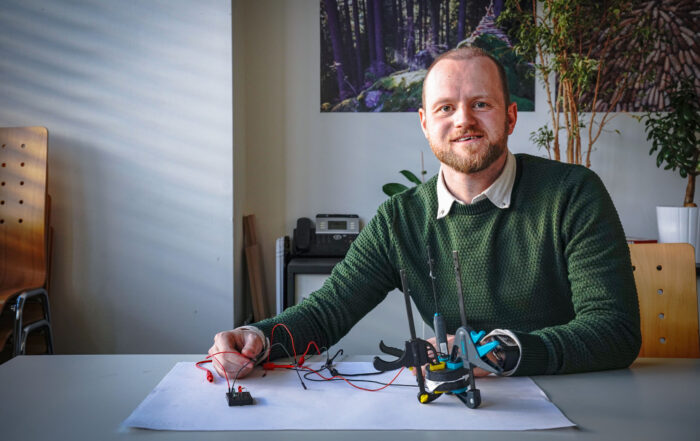 Ein Mann mit Bart, einem grünen Pullover und einem weißen Hemd sitzt an einem Tisch und baut ein elektronisches Projekt mit Drähten und Werkzeugen zusammen. Sonnenlicht wirft Schatten durch den Raum, in dem Pflanzen den Hintergrund schmücken, während er einen Stromspeicher in sein Design integriert. at Hochschule Coburg