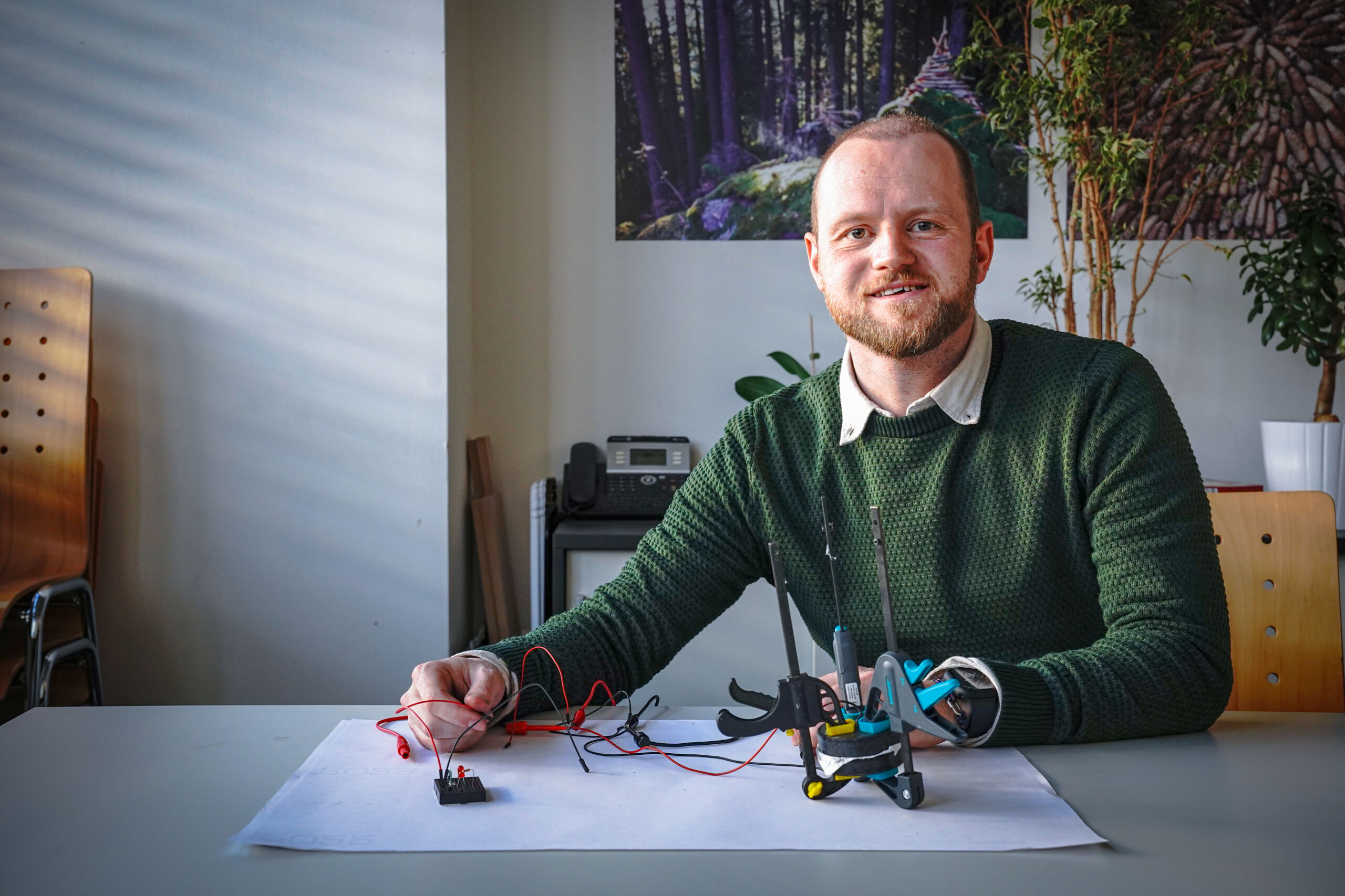 Ein Mann mit Bart, einem grünen Pullover und einem weißen Hemd sitzt an einem Tisch und baut ein elektronisches Projekt mit Drähten und Werkzeugen zusammen. Sonnenlicht wirft Schatten durch den Raum, in dem Pflanzen den Hintergrund schmücken, während er einen Stromspeicher in sein Design integriert. at Hochschule Coburg