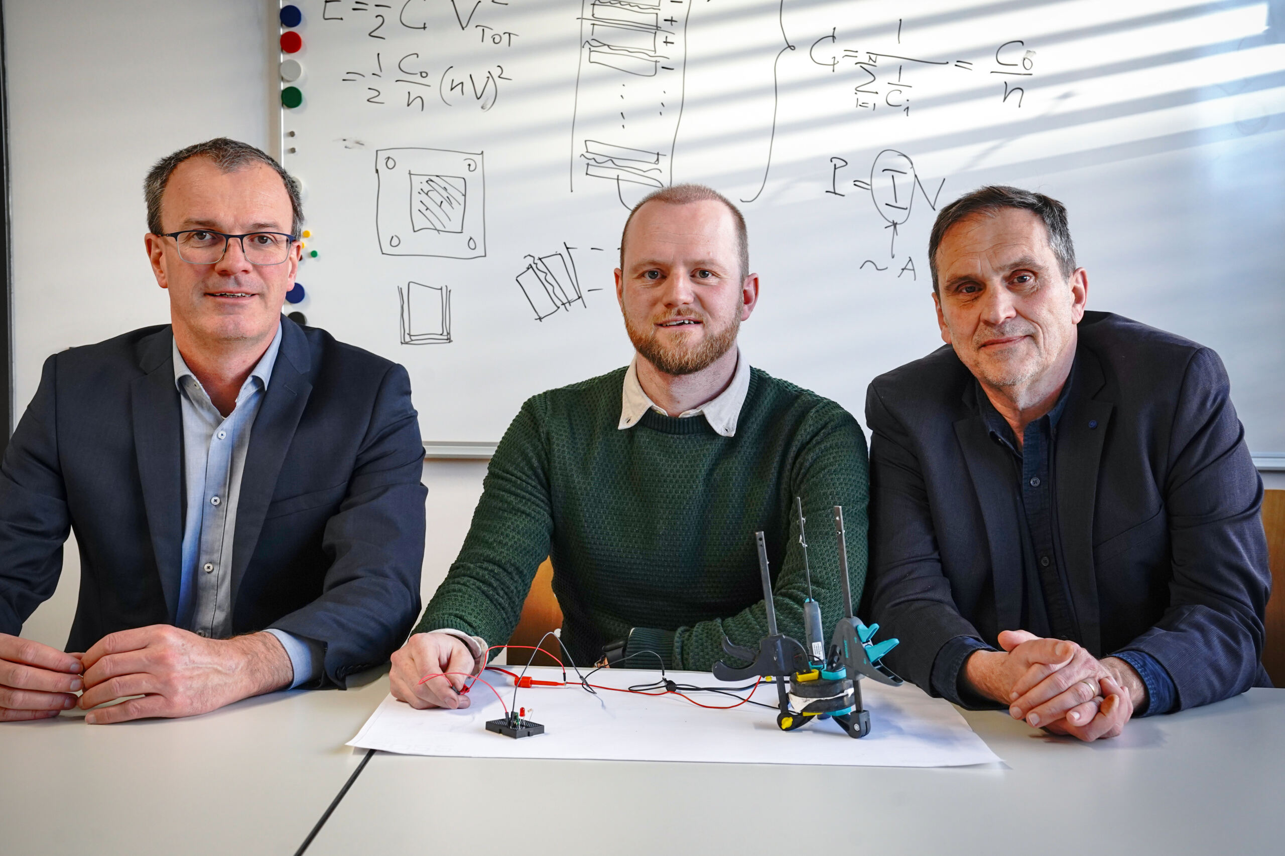 Drei Männer sitzen an einem Tisch mit elektronischen Bauteilen und Diagrammen und diskutieren über Neuerungen bei Stromspeichern. Sie sind in legerer Business-Kleidung gekleidet, im Hintergrund ist ein Whiteboard mit technischen Zeichnungen zu sehen, das ihre konkreten Pläne für die Integration von Technologie und Nachhaltigkeit andeutet. at Hochschule Coburg