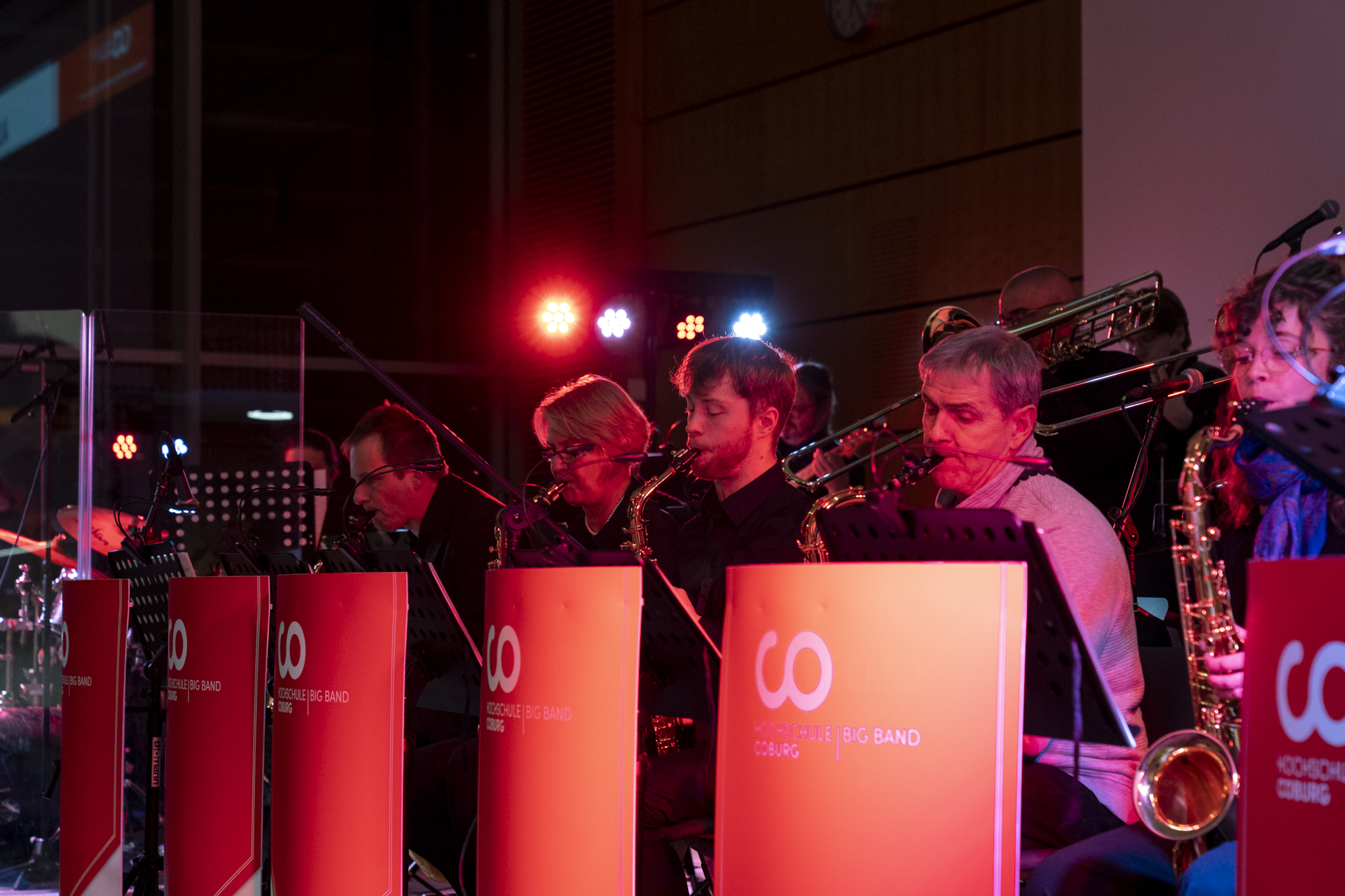 Auf der Bühne tritt eine Big Band auf, die mit Saxophonen und Posaunen das musikalische Flair der 1920er Jahre heraufbeschwört. Sie sitzen hinter roten Notenständern mit dem Logo der Band, während rote und rosa Bühnenlichter eine lebendige Kulisse bilden. at Hochschule Coburg