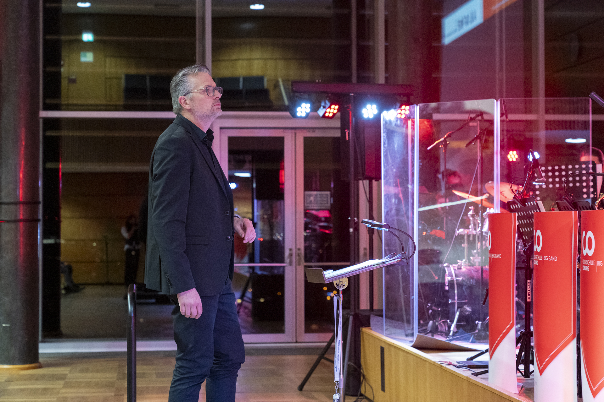 Ein Mann im schwarzen Anzug steht neben einem Podium und blickt auf eine Bühne mit Musikinstrumenten, die an ein Neujahrskonzert der 1920er Jahre erinnern. Rote Lichter erhellen die Szene, und eine Glasbarriere trennt den Mann vom musikalischen Set. at Hochschule Coburg