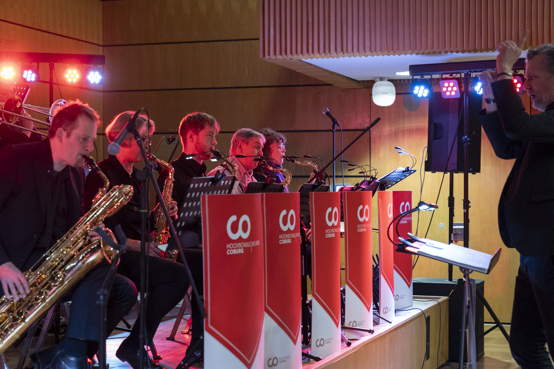 Auf der Bühne tritt eine Jazzband auf, die mit Saxophon- und Trompetenspielern ein Flair der 1920er Jahre verströmt. Rechts steht ein Dirigent, der die Musiker bei diesem musikalischen Fest leitet. Helle Bühnenlichter erhellen die Szene, auf der Notenständer rote Logos mit der Aufschrift „HOCHSCHULZENTRUM COBURG“ tragen. at Hochschule Coburg