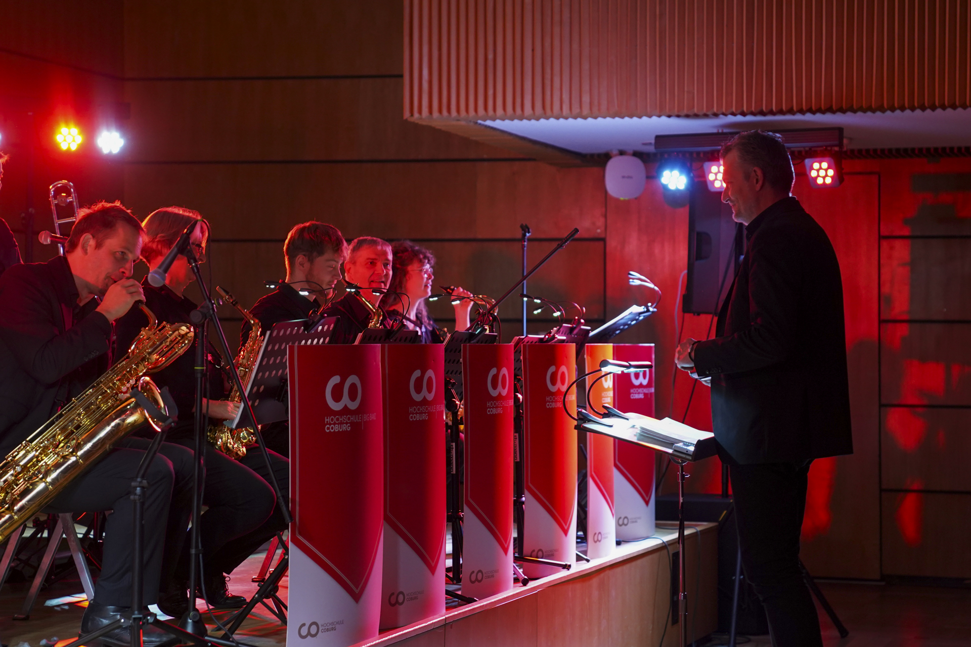 Eine Jazzband verbreitet auf der Bühne eine musikalische Atmosphäre, wobei die Musiker Blechblasinstrumente im Stil der 1920er Jahre spielen. Das Ensemble ist in einer Reihe mit Notenständern aufgestellt und wird von einem Dirigenten im Anzug geleitet, während rotes Licht die Atmosphäre verstärkt und an ein Neujahrskonzert erinnert. at Hochschule Coburg