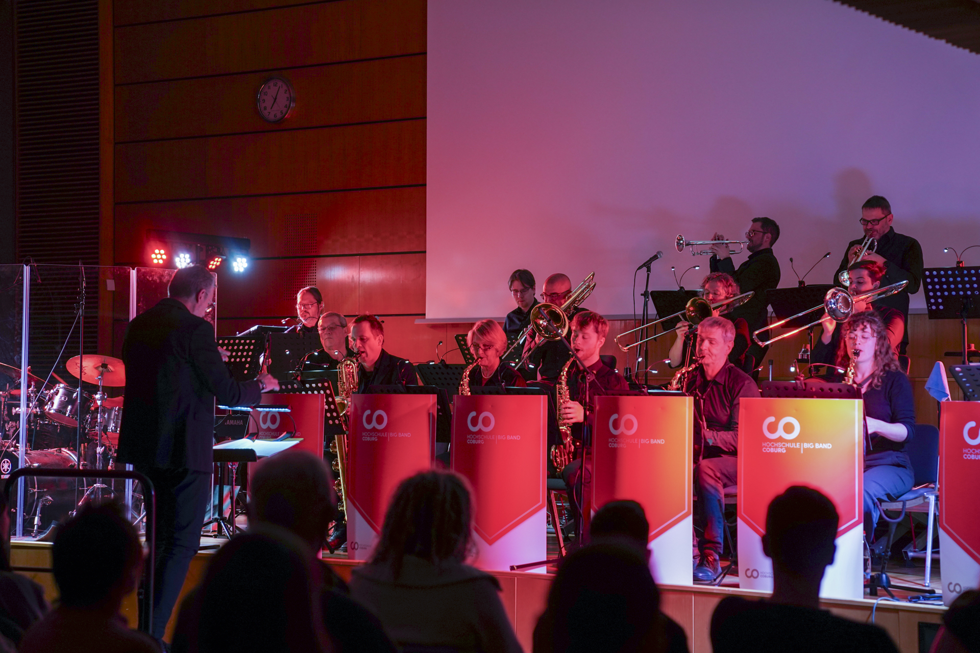 Auf der Bühne spielt eine Big Band unter der Leitung eines Dirigenten, was an ein Neujahrskonzert in den 1920er Jahren erinnert. Musiker spielen Saxophone und Posaunen unter rotem und blauem Licht, während die Uhr an der Wand kurz nach zwölf anzeigt. Das Publikum ist gebannt von diesem musikalischen Spektakel. at Hochschule Coburg