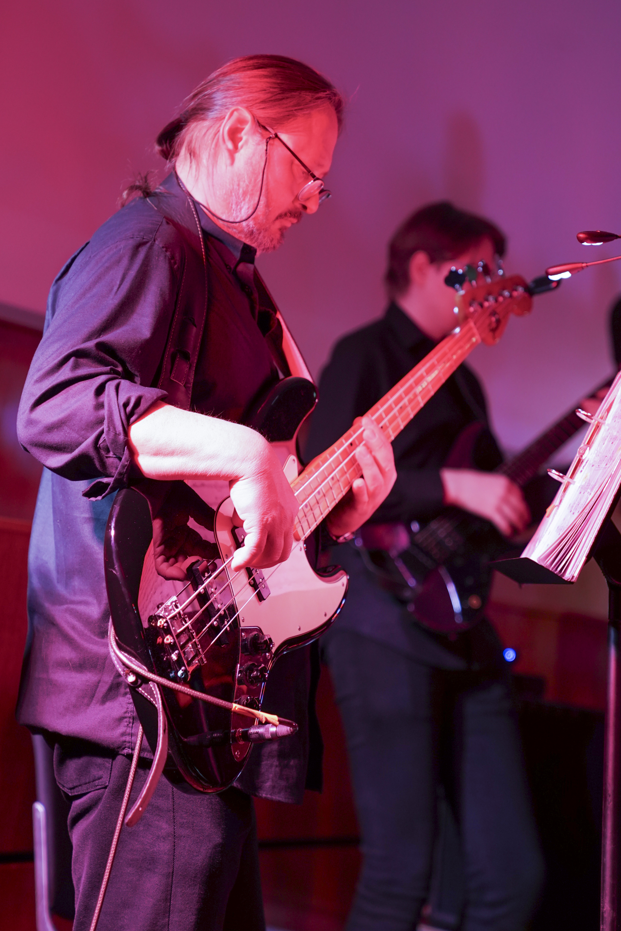 Ein Musiker mit langen Haaren spielt eine E-Bassgitarre und verleiht dem Ganzen einen Hauch von Musikalität, während er Noten liest. Im Hintergrund stimmt ein weiterer Bassist in harmonischen Rhythmus ein. Die Szene, die an ein lebhaftes Neujahrskonzert erinnert, ist in einen rosa Farbton getaucht. at Hochschule Coburg