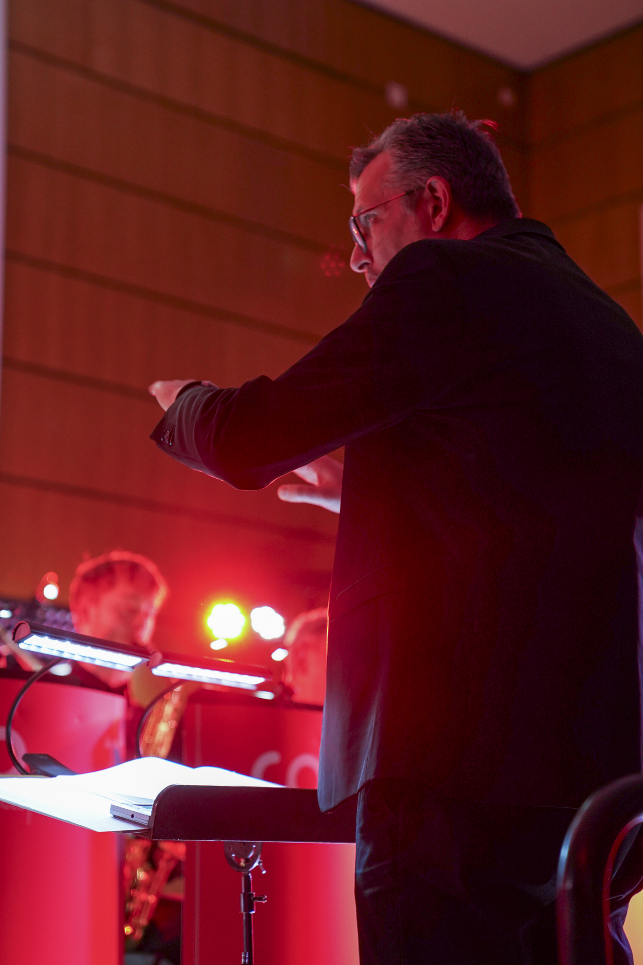 Ein Dirigent im dunklen Anzug leitet ein Orchester beim Neujahrskonzert. Rote und orange Lichter erhellen die Bühne und lassen an die 1920er Jahre denken. Musiker, leicht verschwommen, spielen im Hintergrund Instrumente und schaffen so ein wahrhaft musikalisches Erlebnis. at Hochschule Coburg