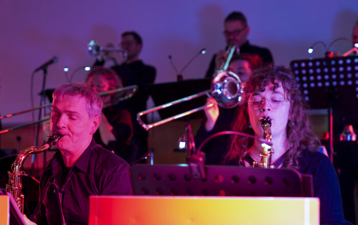 Eine Gruppe von Musikern, die Saxophone und Posaunen spielen, erinnert in einem lebhaften Neujahrskonzert an die 1920er Jahre. Im Vordergrund sind eine Frau und ein Mann mit Saxophonen zu sehen, während im Hintergrund weitere Musiker Posaunen spielen, alles in leuchtendes rotes und blaues Licht getaucht. at Hochschule Coburg