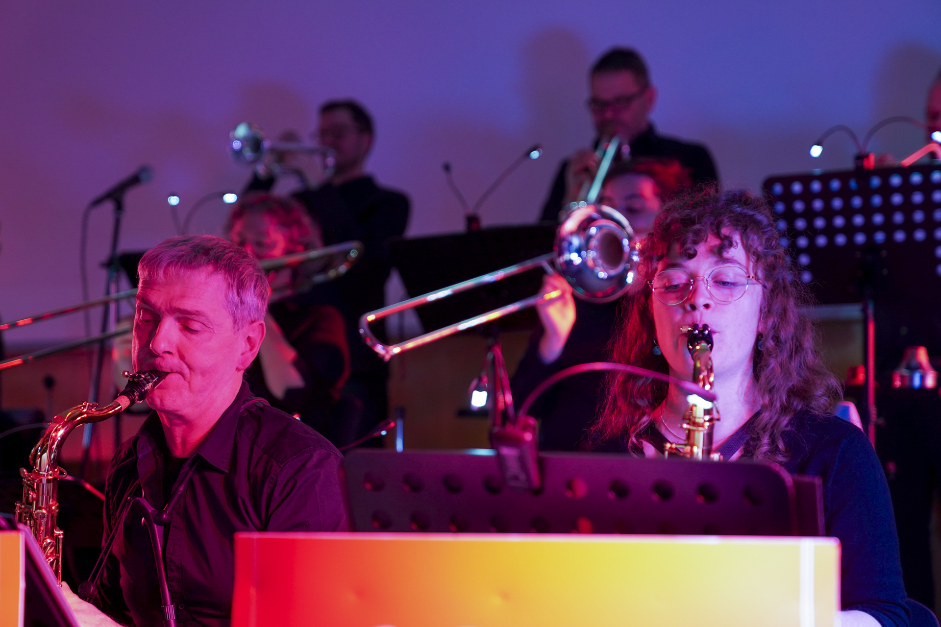 Eine Gruppe von Musikern, die Saxophone und Posaunen spielen, erinnert in einem lebhaften Neujahrskonzert an die 1920er Jahre. Im Vordergrund sind eine Frau und ein Mann mit Saxophonen zu sehen, während im Hintergrund weitere Musiker Posaunen spielen, alles in leuchtendes rotes und blaues Licht getaucht. at Hochschule Coburg