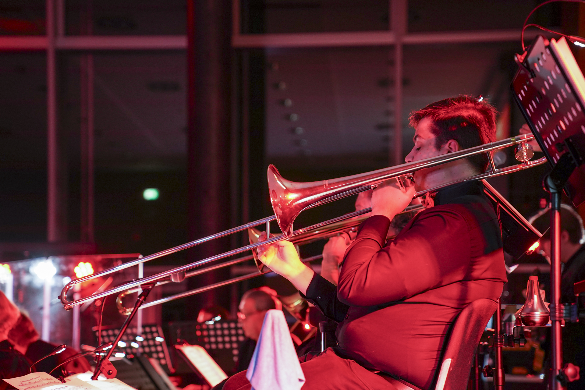 Ein Musiker in einem schwarzen Hemd spielt Posaune in einem Orchestersetup, das an die 1920er Jahre erinnert. Die Szene ist in rotes Licht getaucht und schafft eine eindrucksvolle Kulisse, während andere Musiker mit Notenblättern bereitstehen und sich auf das Neujahrskonzert vorbereiten. at Hochschule Coburg