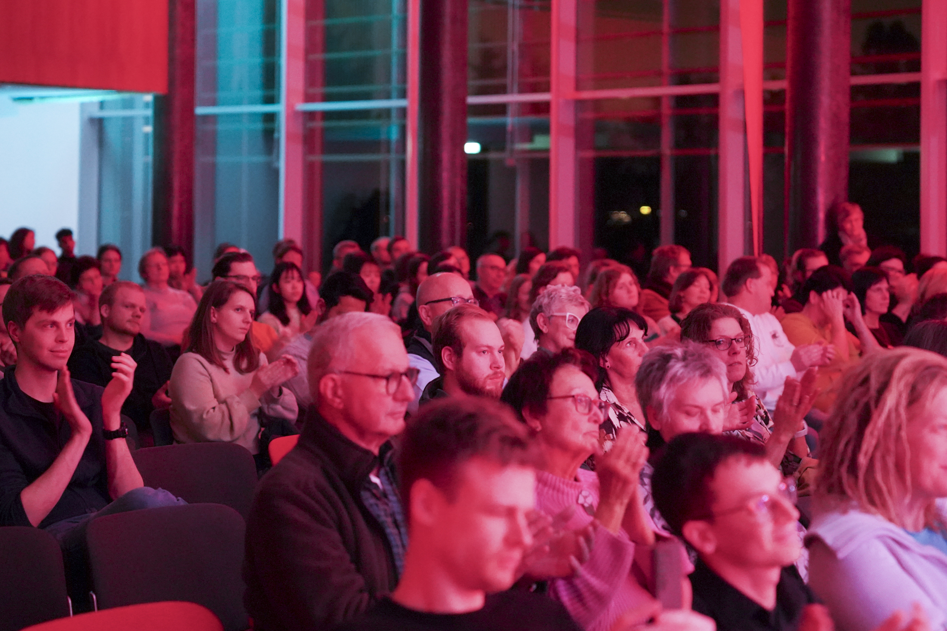 Ein bunt gemischtes Publikum sitzt in Stuhlreihen und verfolgt aufmerksam ein Neujahrskonzert. Der in rotes und blaues Licht getauchte Raum strahlt eine lebendige und fast musikalische Atmosphäre aus, die an die 1920er Jahre erinnert. Einige Leute klatschen, während andere konzentriert auf die Bühne blicken. at Hochschule Coburg