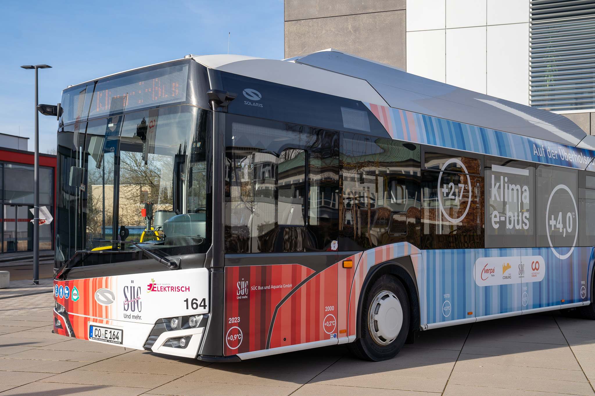 Der Coburger Klima-E-Bus mit seinen bunten Streifen steht vor einem modernen Gebäude. Mit der Aufschrift „klima e-bus“ und der Nummer 164 wirbt dieser Elektrobus inmitten einer glatten gepflasterten Fläche und umgebender Gebäude für Klimaschutz. at Hochschule Coburg