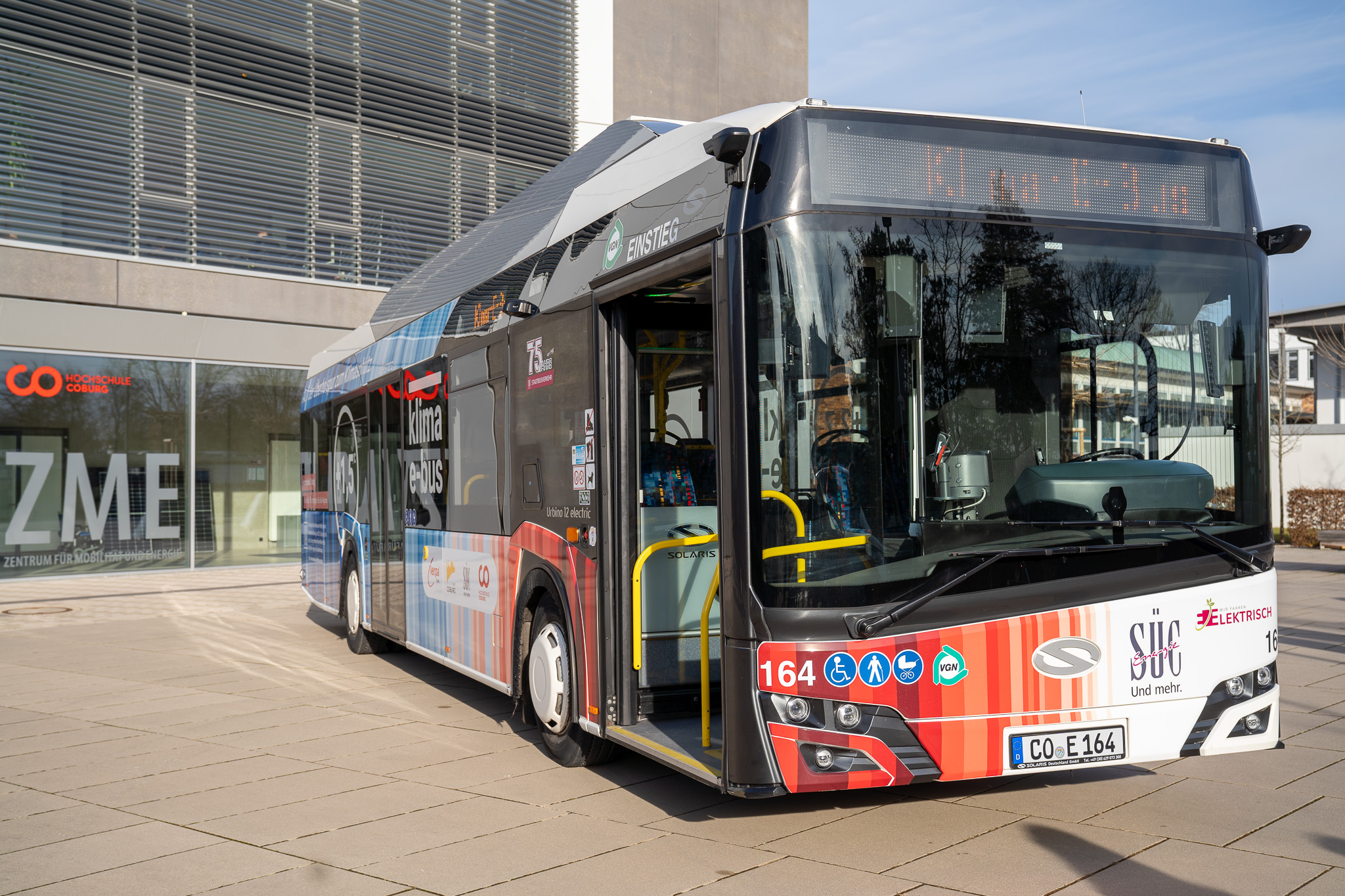 Vor einem Gebäude parkt ein moderner Elektrobus mit farbenfrohem Design. Dieser Coburger Klima-E-Bus, der dem Klimaschutz gewidmet ist, zeigt verschiedene Logos und Schilder. Sein Kennzeichen lautet "CO-E164", im Hintergrund sind die Buchstaben "ZME" zu sehen. at Hochschule Coburg