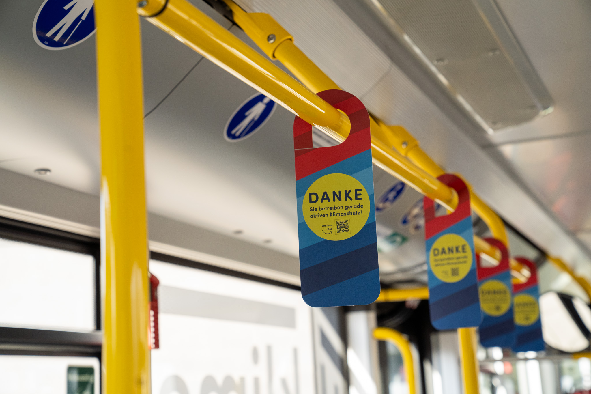 Im Inneren des Coburger E-Busses hängen gelbe Haltestangen und darüber blau-rote Schilder. Auf den Schildern steht in großen Buchstaben „DANKE“ und dankt den Fahrgästen für das Tragen von Masken. An der Decke sind blau-weiße Behindertensymbole zu sehen, die das Engagement der Stadt für den Klimaschutz hervorheben. at Hochschule Coburg