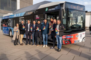 Eine Gruppe von acht Personen steht lächelnd vor einem bunten Stadtbus und hält Auszeichnungsplaketten in den Händen. Der Coburger Klima-E-Bus zeigt verschiedene Grafiken und Texte, die sein Engagement für den Klimaschutz hervorheben. Die Szene ist im Freien, im Hintergrund ist ein Gebäude zu sehen. at Hochschule Coburg