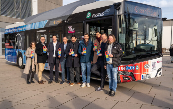 Eine Gruppe von acht Personen steht lächelnd vor einem bunten Stadtbus und hält Auszeichnungsplaketten in den Händen. Der Coburger Klima-E-Bus zeigt verschiedene Grafiken und Texte, die sein Engagement für den Klimaschutz hervorheben. Die Szene ist im Freien, im Hintergrund ist ein Gebäude zu sehen. at Hochschule Coburg