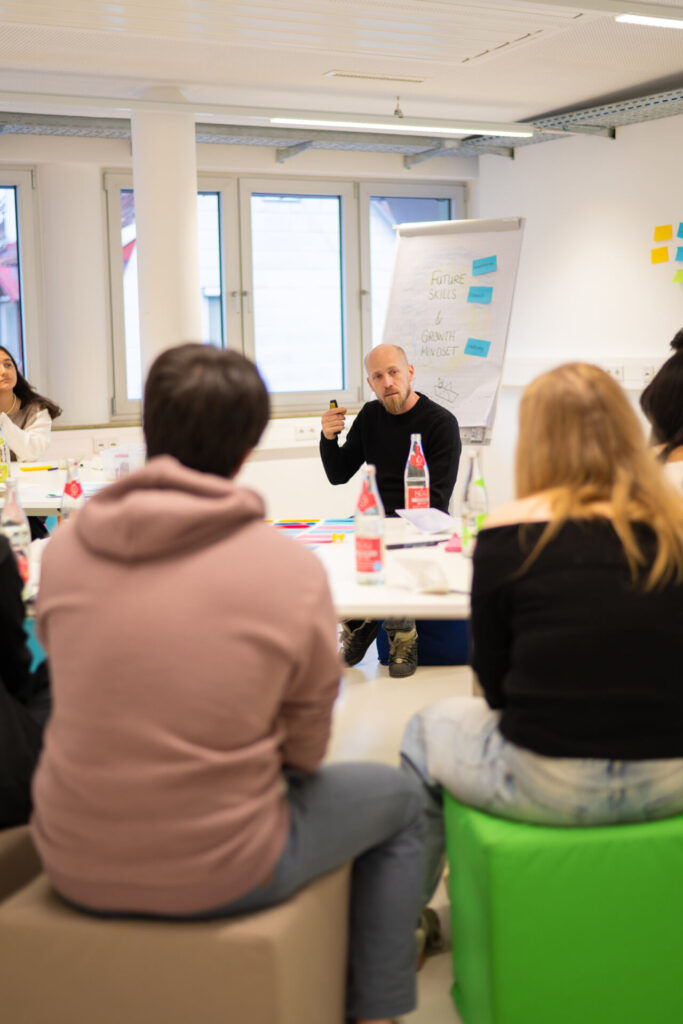 Ein Mann sitzt und gestikuliert, während er zu einer Gruppe um einen Tisch in einem hell erleuchteten Raum spricht. Die Gruppe ist aufmerksam, auf dem Tisch stehen Notizbücher und Getränke. Im Hintergrund ist ein Flipchart mit Notizen darauf. at Hochschule Coburg