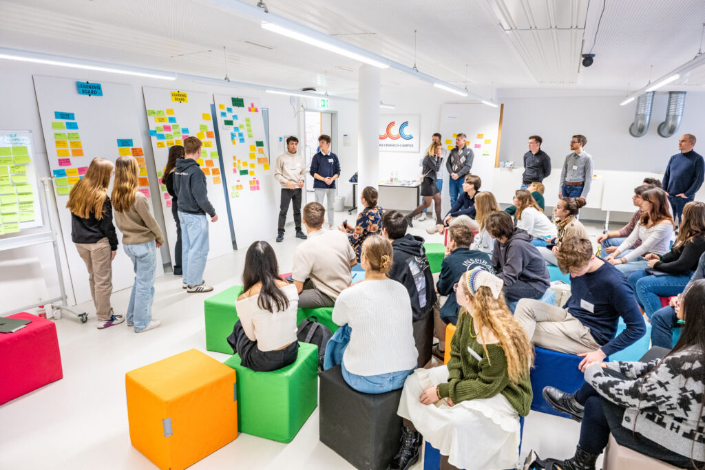 Eine Gruppe von Menschen hat sich in einem hellen Raum zu einem Workshop versammelt. Einige Personen stehen und präsentieren vor Whiteboards, die mit bunten Haftnotizen bedeckt sind, während andere auf Sitzwürfeln sitzen und aufmerksam zuhören. at Hochschule Coburg