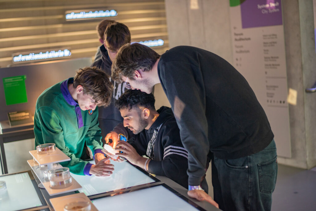 Vier Personen nehmen an einer wissenschaftlichen Ausstellung teil. Eine Person untersucht ein Objekt mit einer Lupe, während sich andere um sie herum versammeln und beobachten. Sie wirken konzentriert und die Kulisse ist eine interaktive Museumsausstellung. at Hochschule Coburg