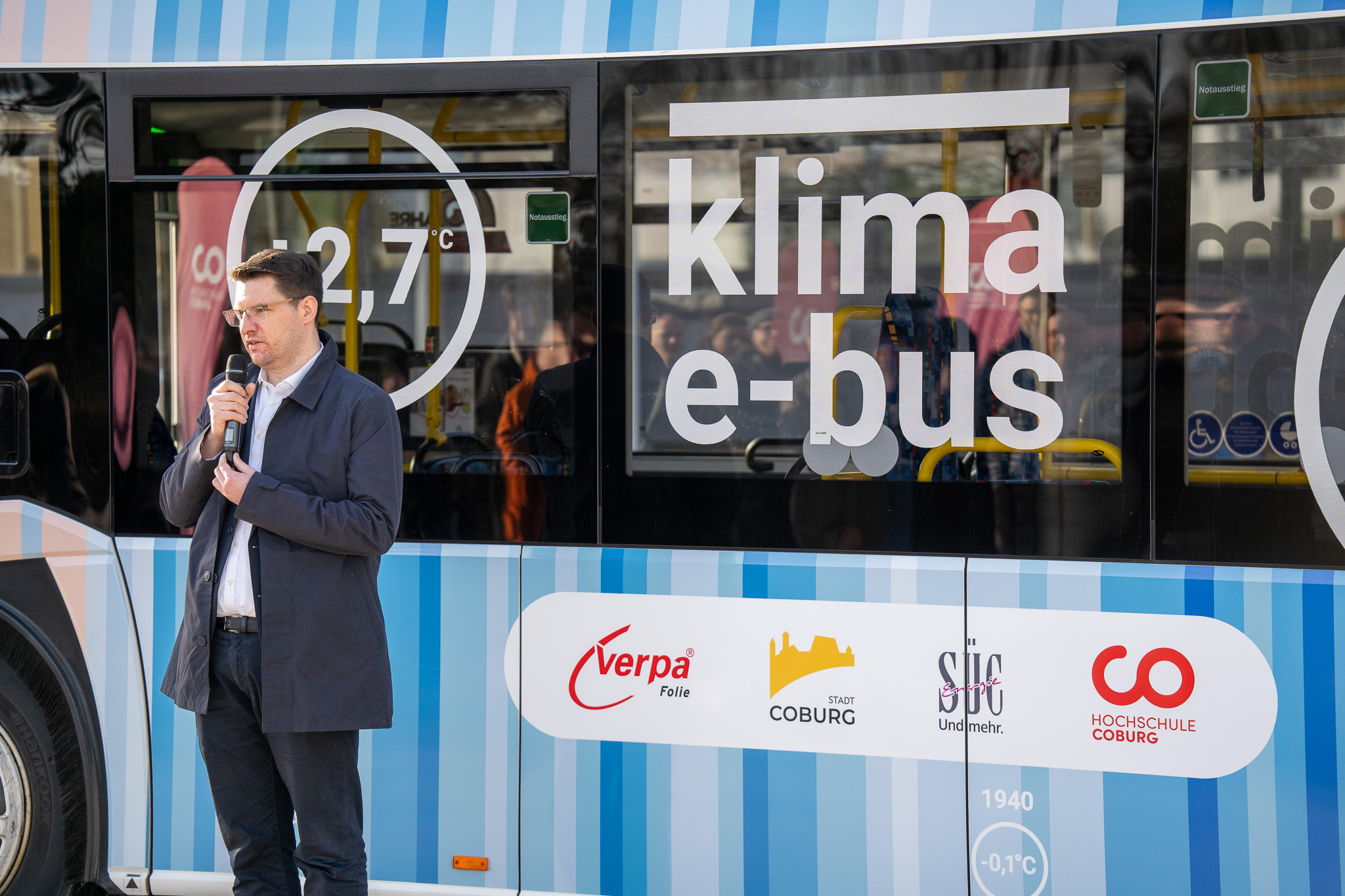 Eine Person spricht in ein Mikrofon neben dem innovativen Klima-E-Bus, der stolz die Marken von Verpa, Stadt Coburg und Hochschule Coburg präsentiert. Im Bus ist eine Gruppe von Menschen zu sehen, die den Geist des Klimaschutzes verkörpern. at Hochschule Coburg