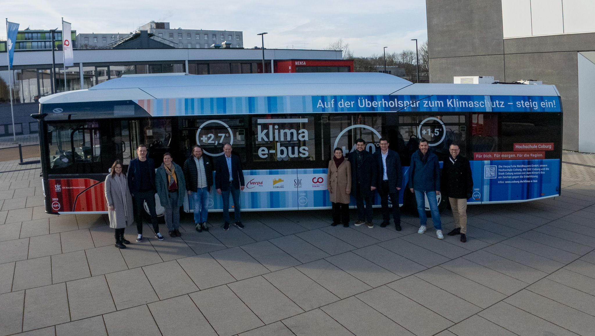 Eine Gruppe von Menschen steht in Coburg vor einem „Klima-E-Bus“, der Klimaslogans und Temperatursymbole trägt. Der Bus, der die Klimaschutzbemühungen symbolisiert, steht auf einer gepflasterten Fläche, im Hintergrund sind Gebäude und ein Schild zu sehen. at Hochschule Coburg