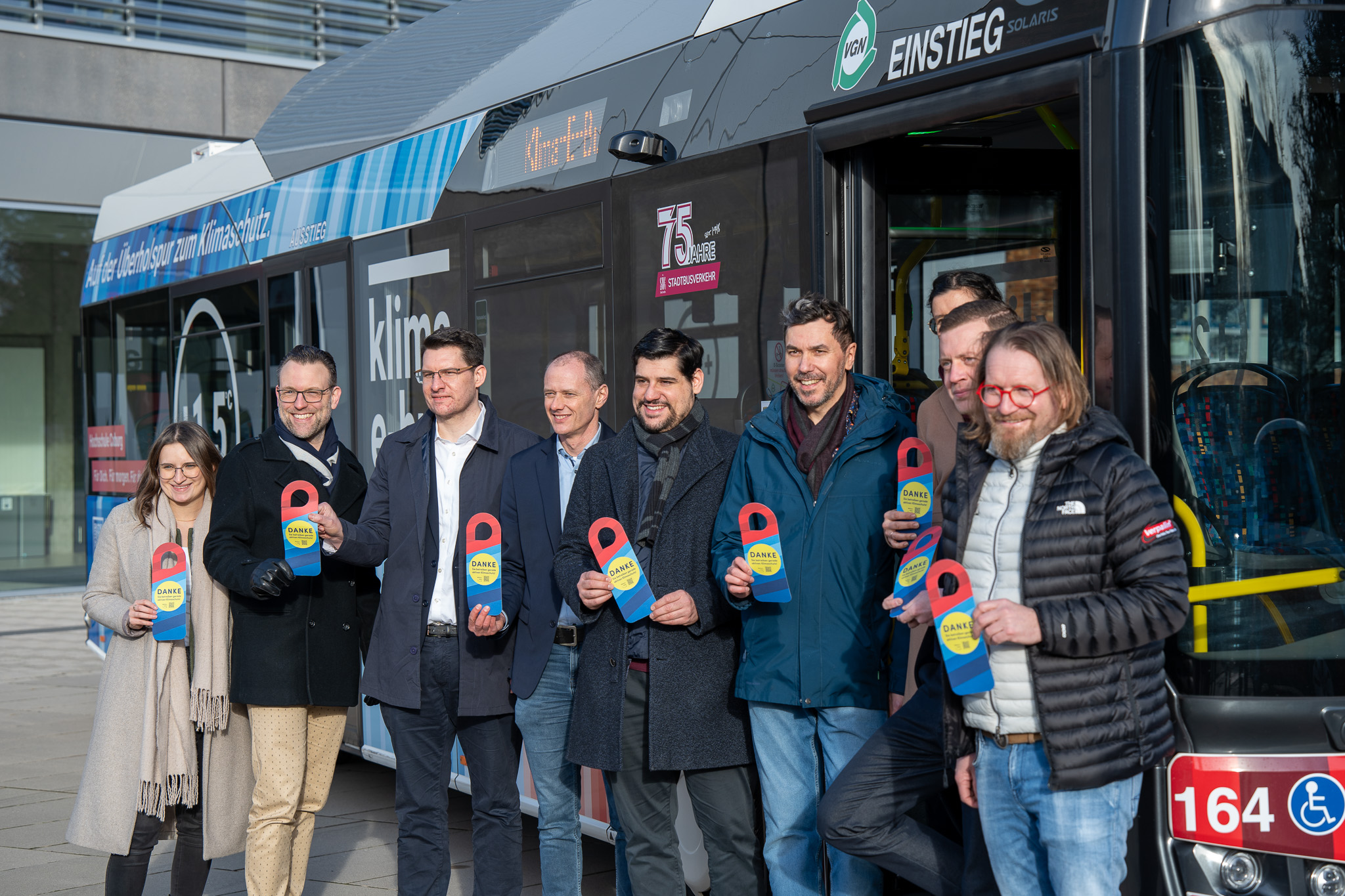 Eine Gruppe von Menschen steht vor einem Klima-E-Bus und hält bunte Gegenstände in Form von Schlüsselanhängern in den Händen. Auf der Seite des Busses steht der Schriftzug „Klimaschutz“. Sie tragen Winterkleidung und die Szene spielt im Freien in einem städtischen Gebiet. at Hochschule Coburg