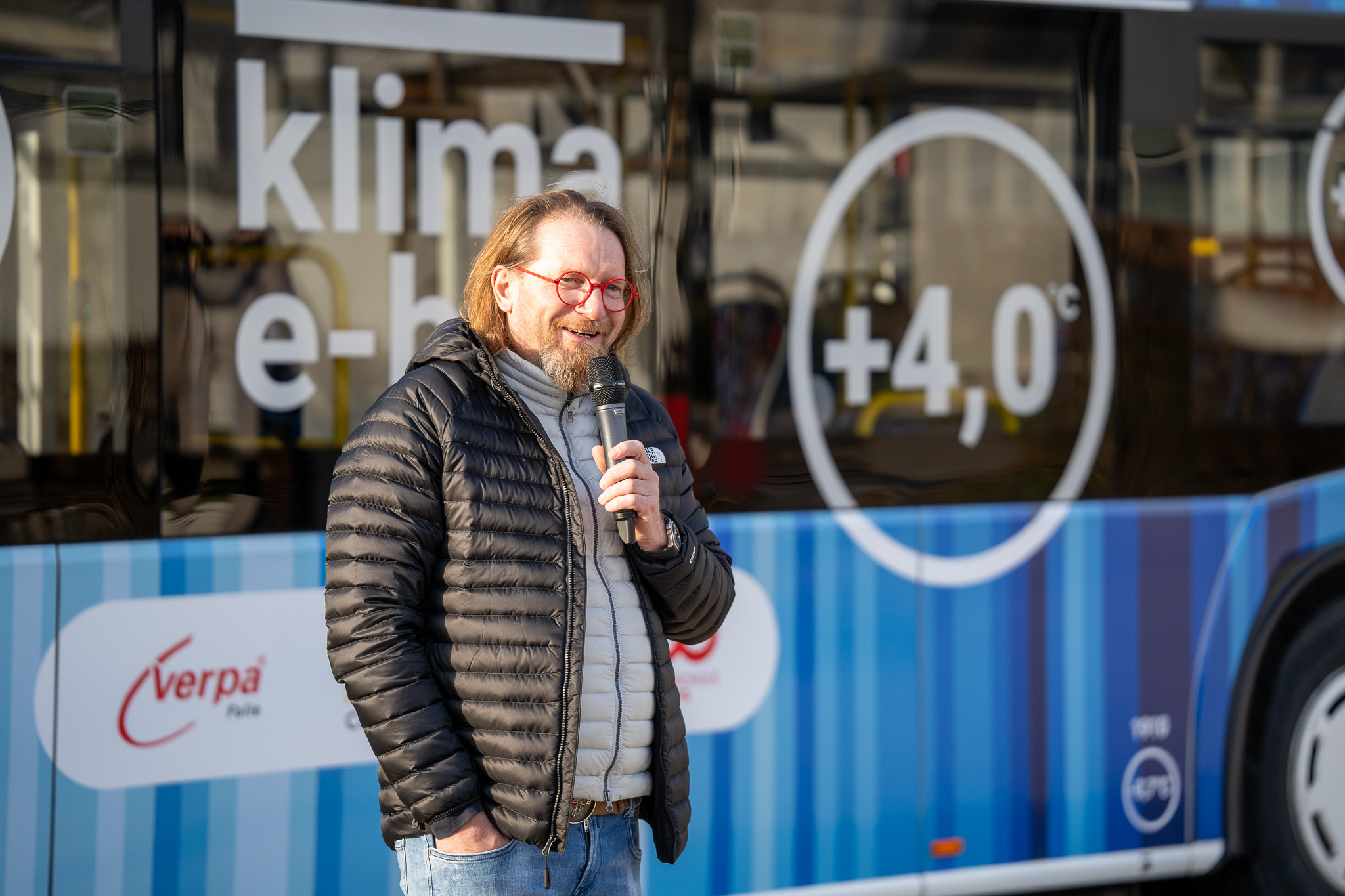 Eine Person mit langen Haaren und Brille, bekleidet mit einer schwarzen Pufferjacke, hält ein Mikrofon und steht vor dem Coburger Klima-E-Bus, der mit blauen Streifen und der Aufschrift „Klima E-Bus +4,0°C“ geschmückt ist und auf die Bemühungen zum Klimaschutz aufmerksam macht. at Hochschule Coburg