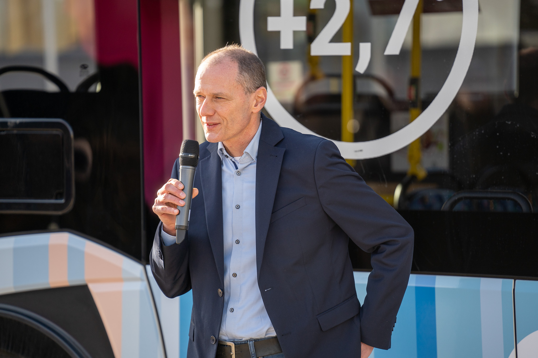 Ein Mann in blauem Hemd und dunklem Blazer hält ein Mikrofon und spricht leidenschaftlich über Klimaschutz vor dem Klima-E-Bus. Im Hintergrund ist ein Teil eines Busdesigns mit den Zahlen „+2,7“ zu sehen. at Hochschule Coburg