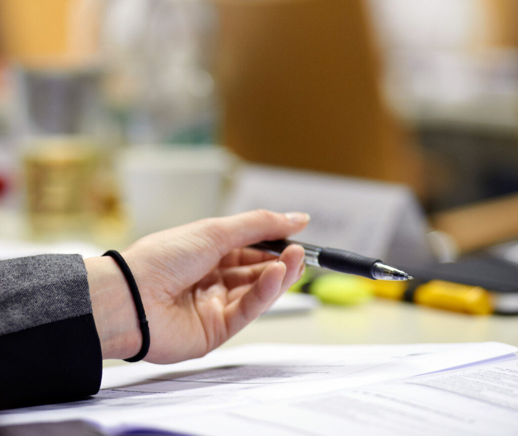 Eine Hand hält einen Stift über verstreuten Dokumenten auf einem Schreibtisch. Die Person trägt eine dunkle Jacke und ein schwarzes Haargummi um das Handgelenk. Der Hintergrund ist verschwommen, verschiedene Büromaterialien sind sichtbar. at Hochschule Coburg