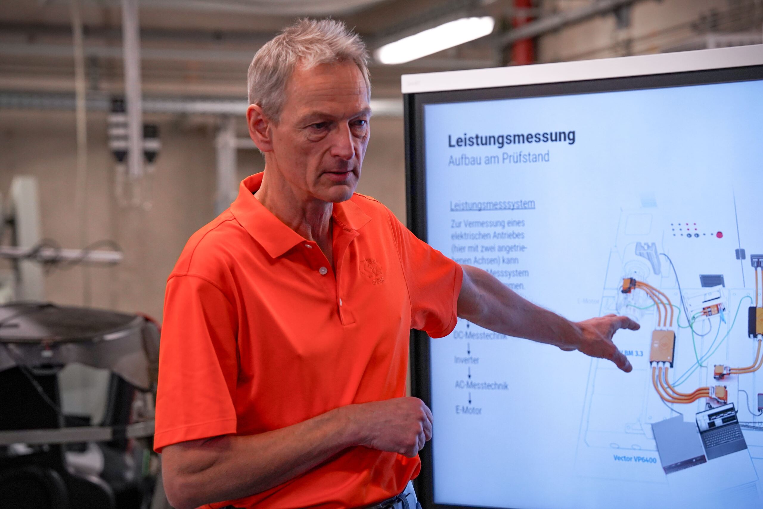 Ein Mann in einem orangefarbenen Hemd erklärt in einem technischen Umfeld ein Diagramm auf einem großen Bildschirm, vermutlich Teil eines E-Auto-Programms. Der Bildschirm zeigt ein Schema mit der Aufschrift „Leistungsmessung“ mit verschiedenen Komponenten und Leitungen, die für Schulungen zur Kfz-Ausbildung wichtig sind. at Hochschule Coburg