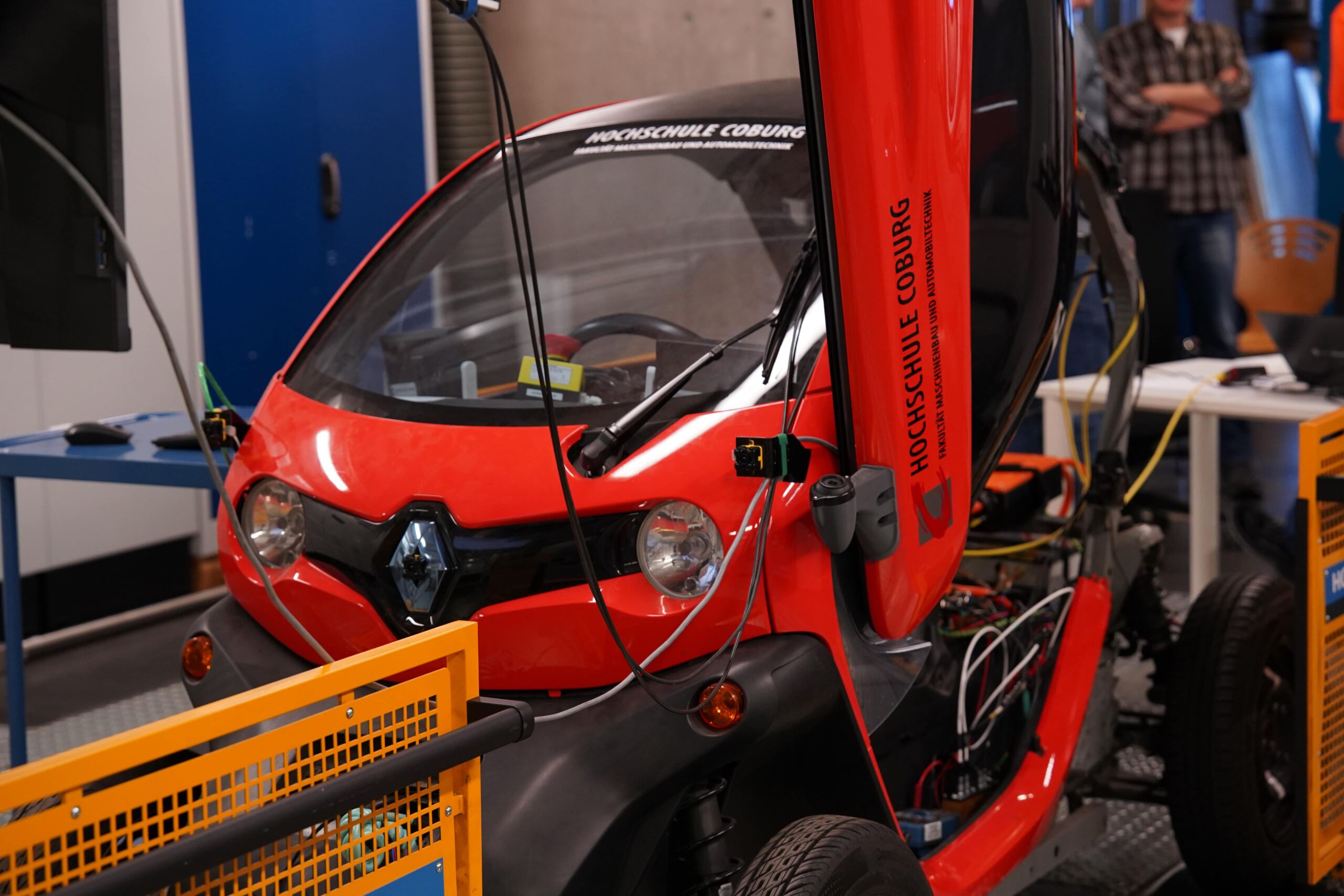 Ein rotes E-Auto mit freiliegenden Kabeln wird im Innenbereich ausgestellt. Die Motorhaube ist teilweise geöffnet und zeigt die inneren Komponenten des Fahrzeugs. Auf der Seite steht „Hochschule Coburg“, was ein Zentrum für Kfz-Ausbildung symbolisiert. Im Hintergrund sind Menschen und Ausrüstung zu sehen. at Hochschule Coburg