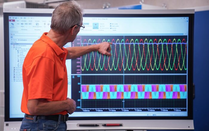 Eine Person in einem orangefarbenen Hemd zeigt auf einen großen Bildschirm, auf dem bunte Sinuskurven und Datendiagramme angezeigt werden, was auf eine Bildungsumgebung mit dem Schwerpunkt auf Weiterbildung oder möglicherweise E-Auto-Technologie hindeutet. at Hochschule Coburg