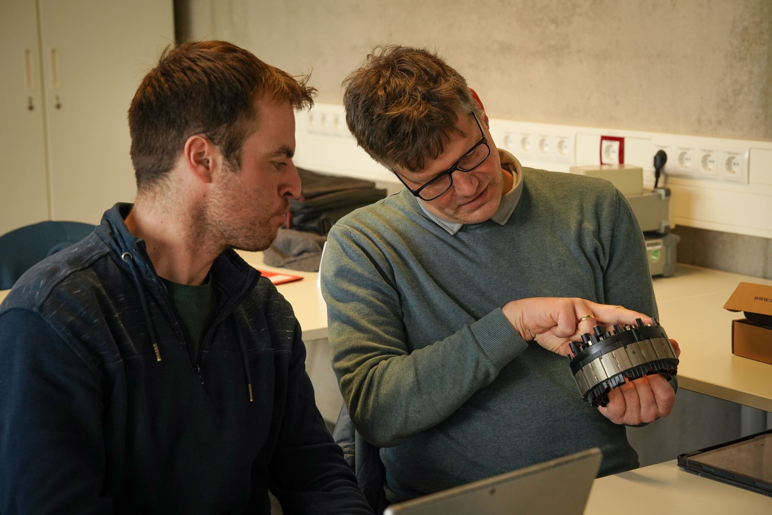 Zwei Männer sitzen an einem Tisch und untersuchen ein mechanisches Bauteil im Zusammenhang mit der Kfz-Ausbildung. Ein Mann mit Brille hält das Teil und zeigt darauf. Sie scheinen sich in einer Werkstatt oder einem Klassenzimmer zu befinden. Im Hintergrund sind Werkzeuge und Steckdosen zu sehen. at Hochschule Coburg