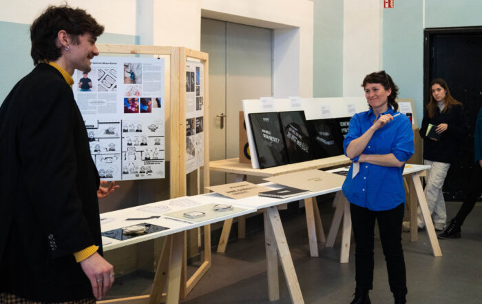 Eine Gruppe von Menschen beobachtet in einer Galerie einen Mann, der seine Arbeit präsentiert. In der ungezwungenen Atmosphäre wird über dieses Studierenden-Projekt diskutiert, in dem Architekturzeichnungen und Fotografien gezeigt werden, die Design-Aktivismus mit kreativer Vision verbinden. at Hochschule Coburg