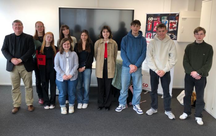 Eine Gruppe von zehn Personen steht drinnen und posiert für ein Foto vor einem großen Fernsehbildschirm mit einem Plakat und einem Banner im Hintergrund. at Hochschule Coburg