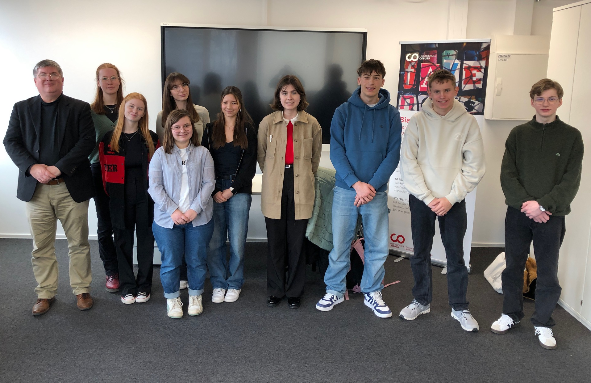 Eine Gruppe von zehn Personen steht drinnen und posiert für ein Foto vor einem großen Fernsehbildschirm mit einem Plakat und einem Banner im Hintergrund. at Hochschule Coburg