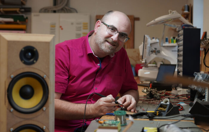 Eine Person in rotem Hemd und Brille lächelt und hält einen Lötkolben an einer Werkbank im CREAPOLIS Makerspace. Inmitten des Prototyping-Rummels liegen elektronische Komponenten und Werkzeuge verstreut auf dem Tisch, links steht ein großer Lautsprecher. at Hochschule Coburg