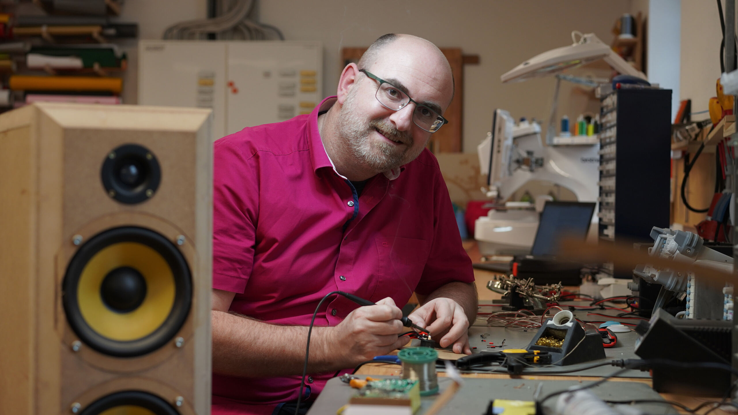 Eine Person in rotem Hemd und Brille lächelt und hält einen Lötkolben an einer Werkbank im CREAPOLIS Makerspace. Inmitten des Prototyping-Rummels liegen elektronische Komponenten und Werkzeuge verstreut auf dem Tisch, links steht ein großer Lautsprecher. at Hochschule Coburg