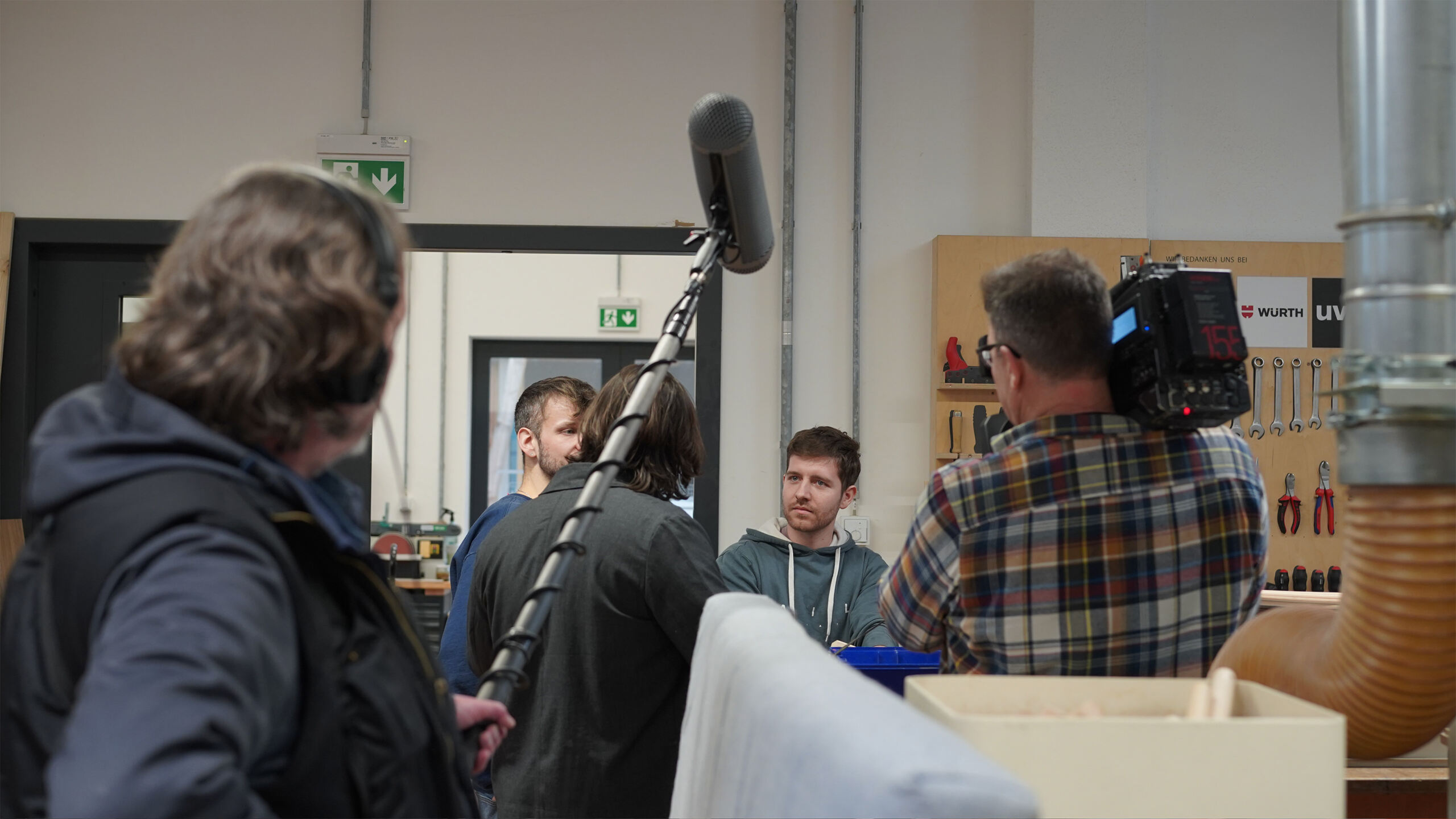 Ein Filmteam filmt eine Szene im Innenbereich des CREAPOLIS Makerspace. Eine Person hält ein Galgenmikrofon, während eine andere eine Videokamera bedient. Zwei Personen stehen vor einem Hintergrund, an dessen Wand Prototyping-Tools hängen, und fangen die Essenz handwerklicher Kreativität ein. at Hochschule Coburg