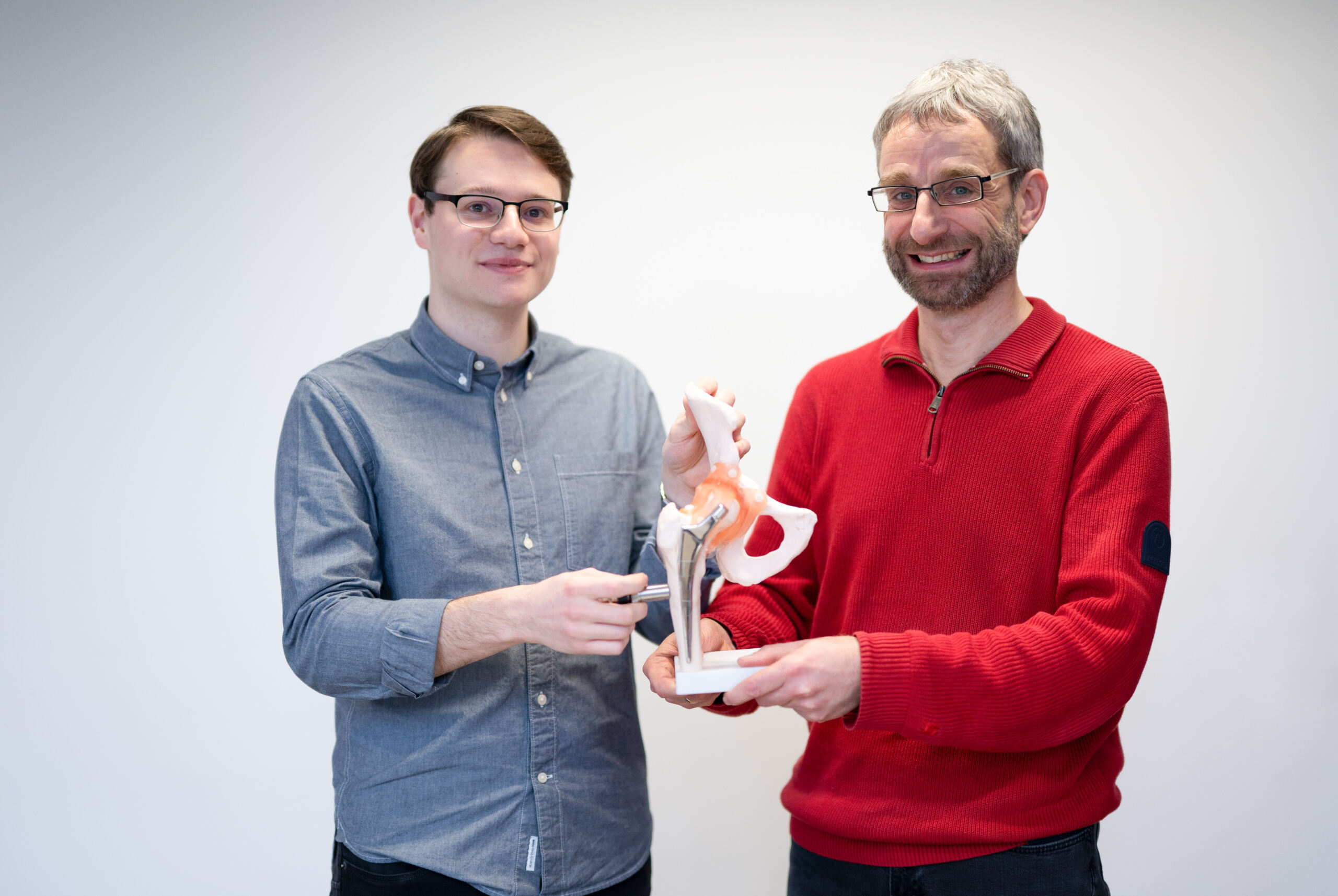 Zwei Männer lächeln und halten ein 3D-Modell eines Beckengelenks in der Hand. Der Mann links im grauen Hemd und der Mann rechts im roten Pullover präsentieren ihre Forschung zu Hüftprothesen vor einem schlichten weißen Hintergrund. at Hochschule Coburg